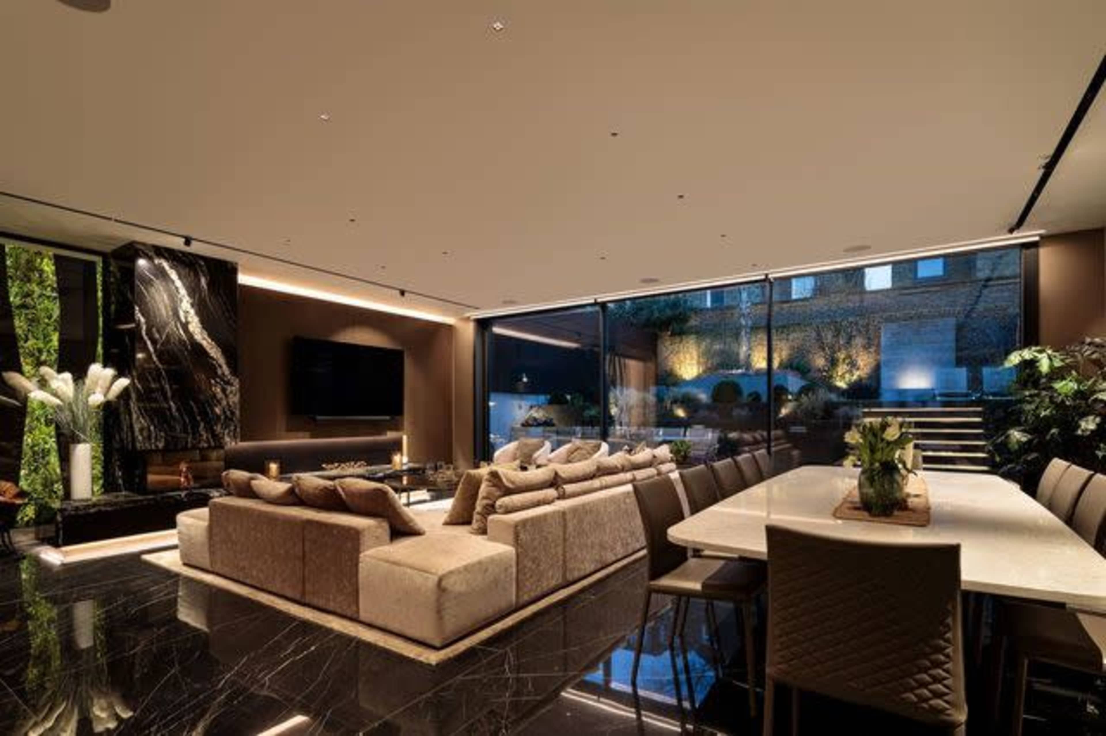 A modern living room features a large sectional sofa, a dining table, and floor-to-ceiling windows overlooking an outdoor space.