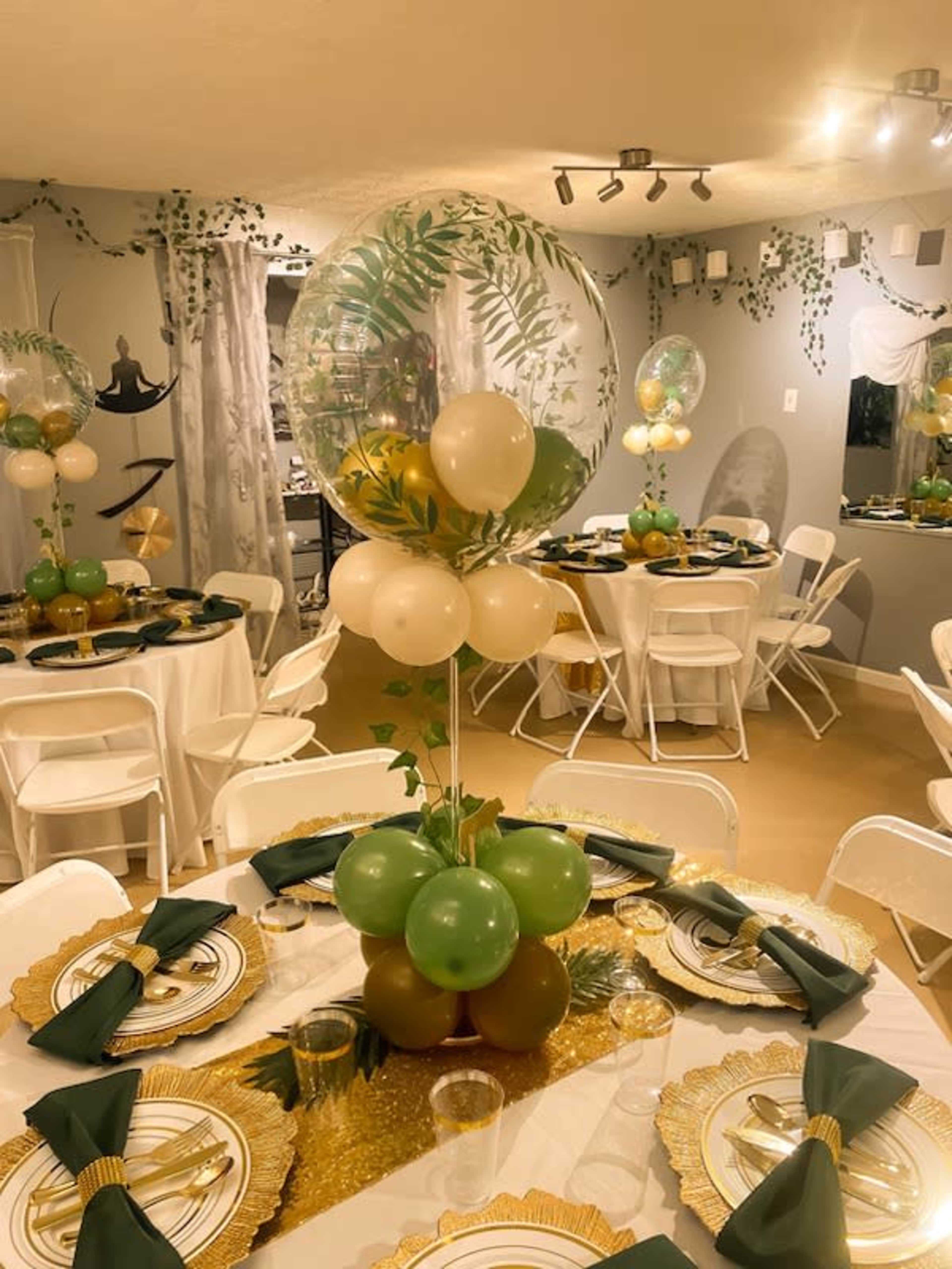 The image shows a decorated event space featuring tables set with golden plates, green and gold balloons, and white chairs.