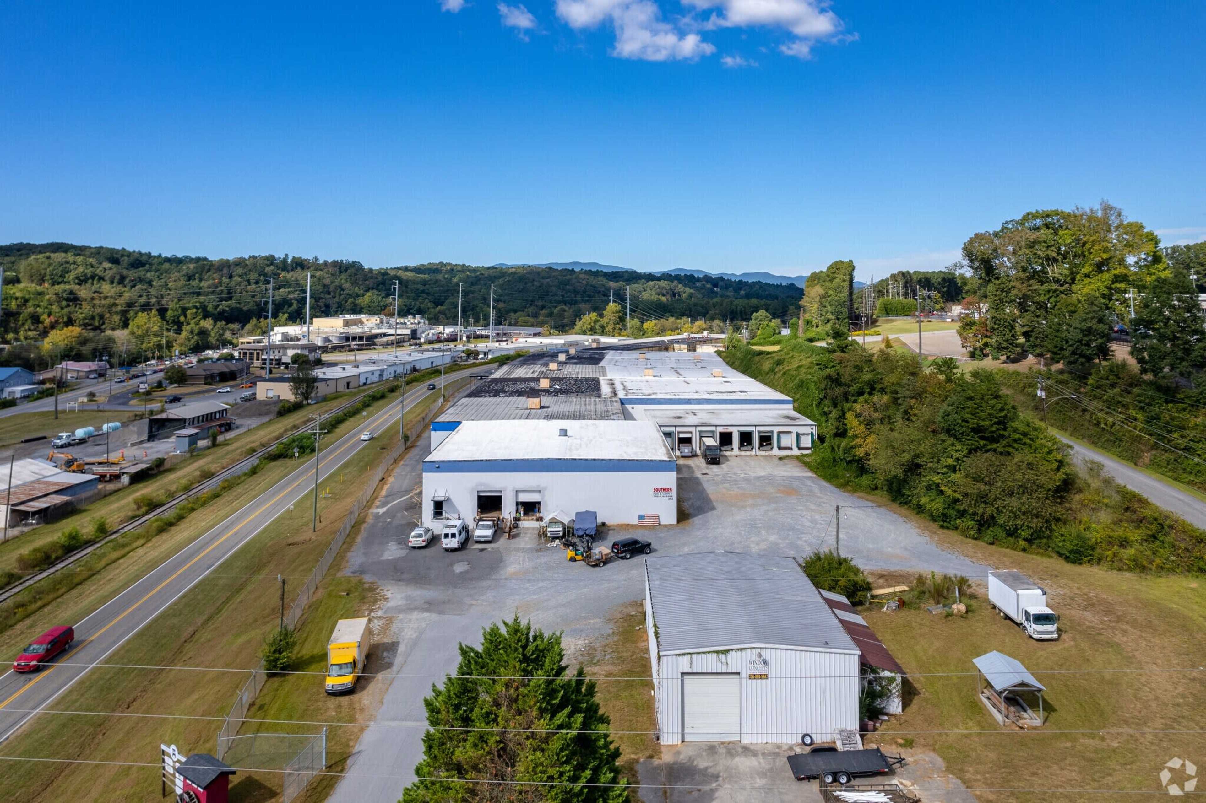 A large industrial complex with multiple warehouse buildings along a road and surrounded by trees and hills in the background.