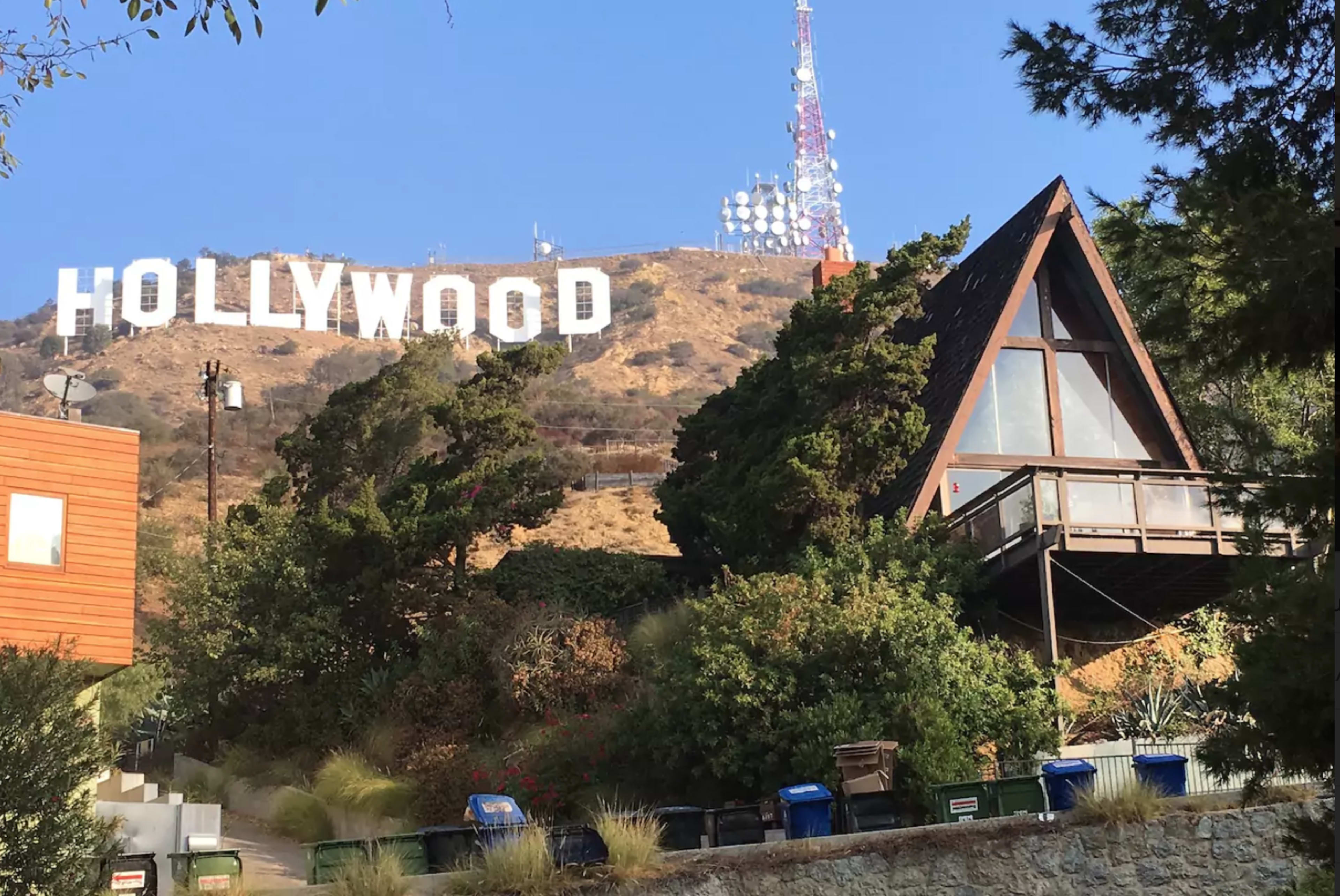 The iconic Hollywood sign is prominently displayed on a hillside above a modern A-frame house surrounded by greenery.