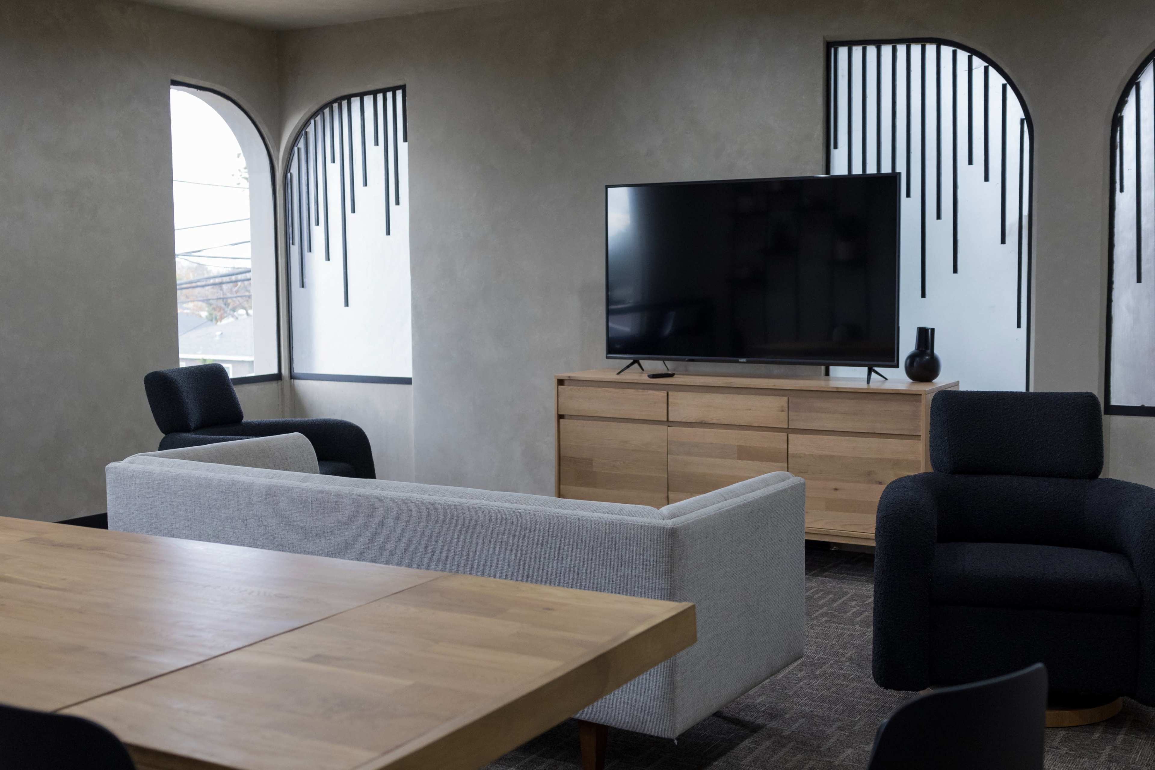 A modern lounge area features a large television on a wooden cabinet, with a gray sofa, two black armchairs, and a wooden dining table.