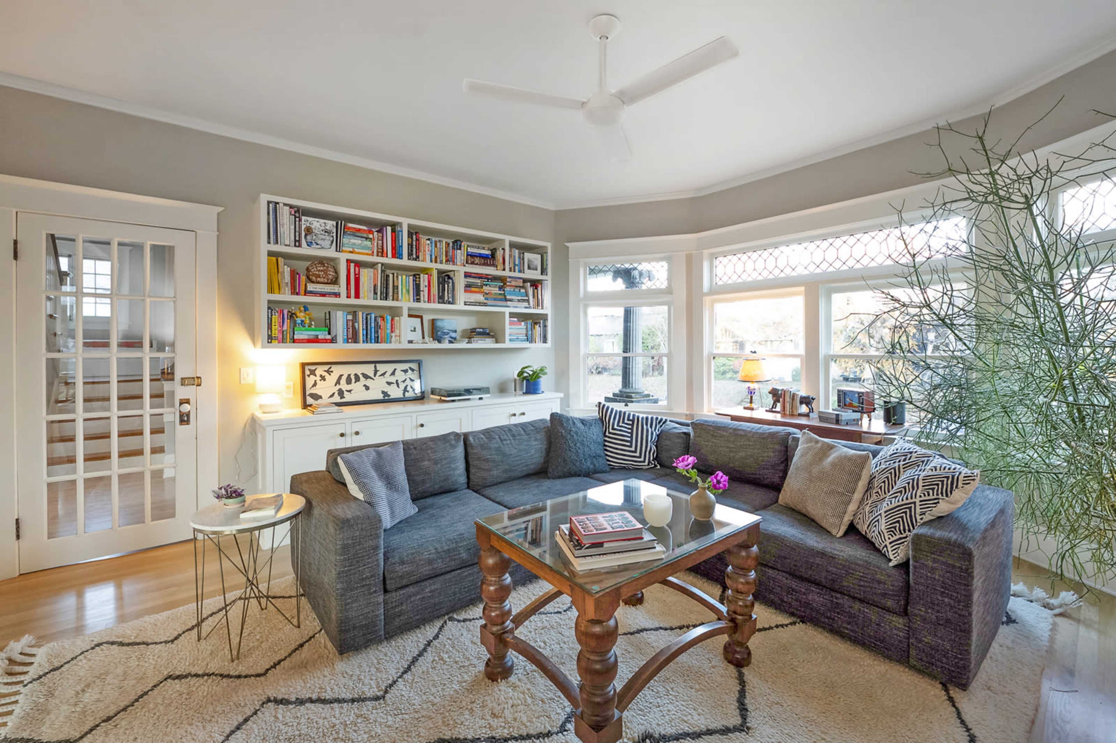 A modern living room features a large sectional sofa, a glass coffee table, built-in bookshelves filled with books, and natural light streaming through multiple windows.