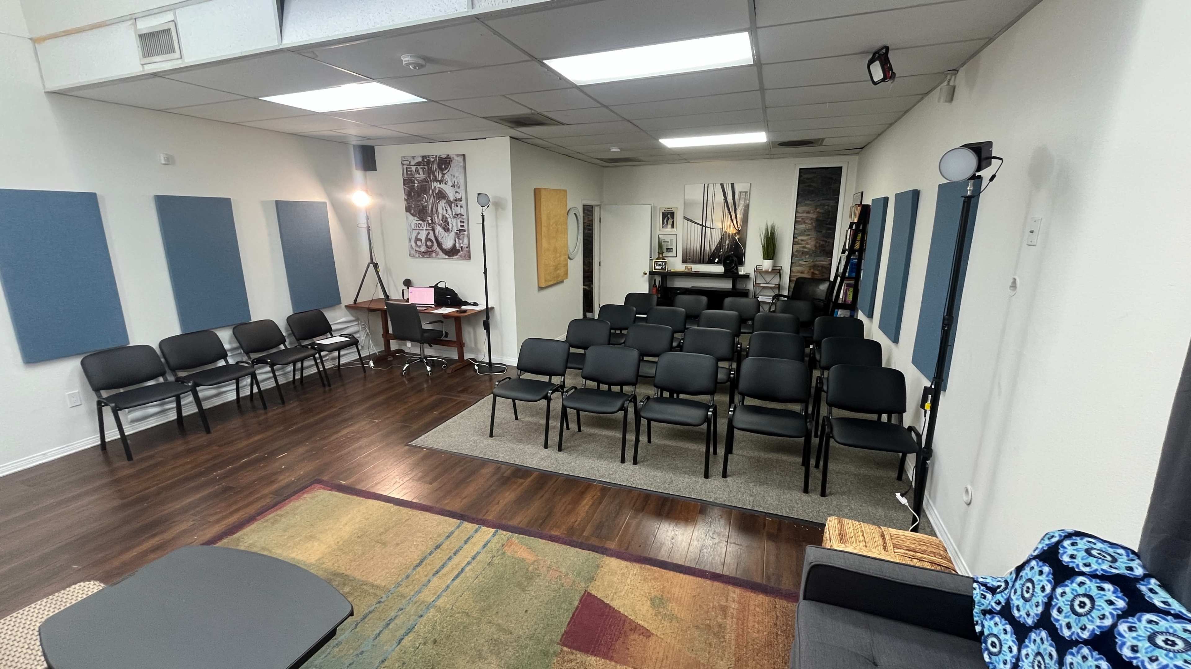 The image shows a room set up for an event, with rows of black chairs facing a table and a backdrop featuring wall decorations and lighting equipment.