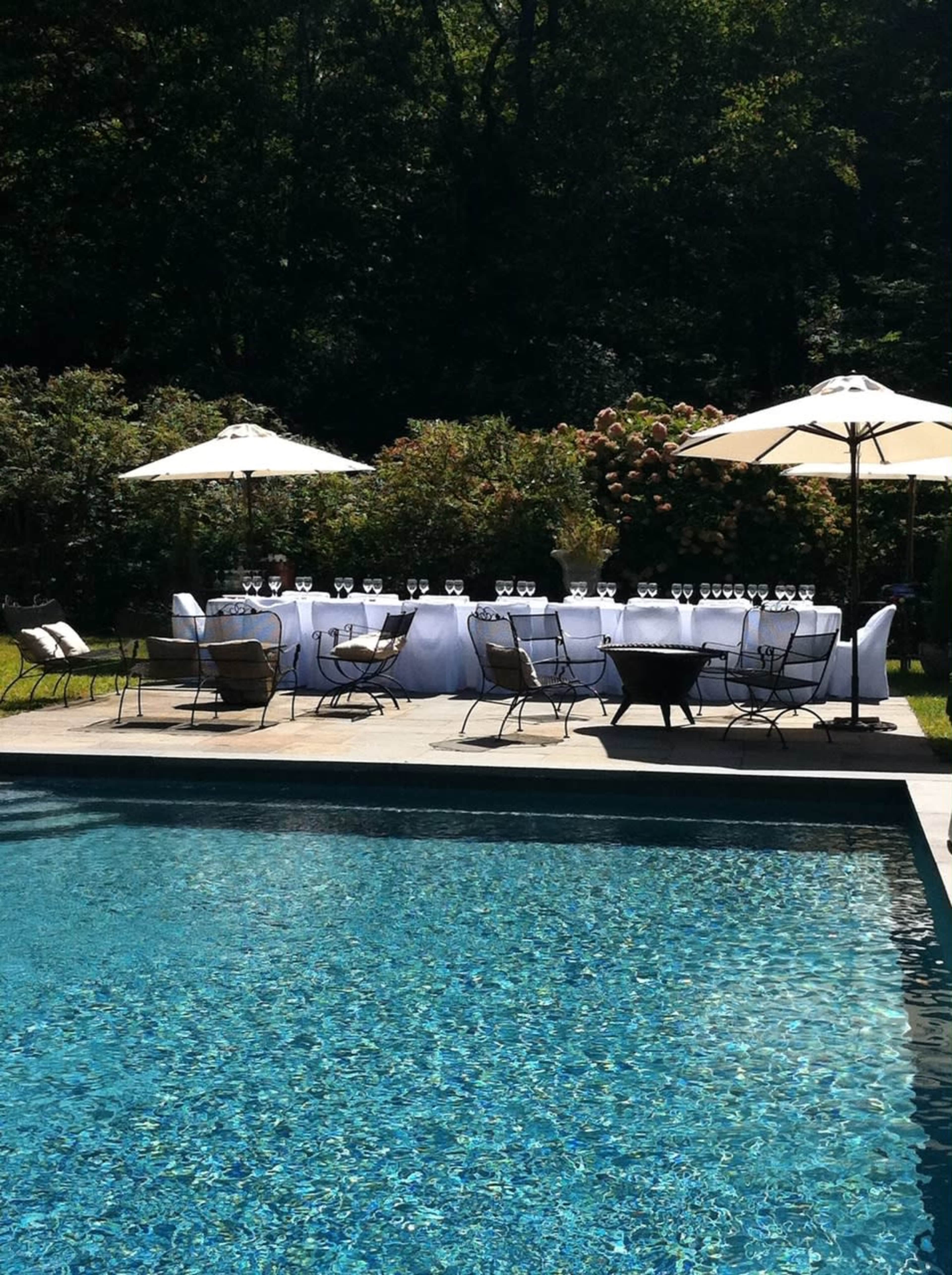 A long table set for dining with glasses sits beside a swimming pool and under umbrellas, surrounded by green foliage.