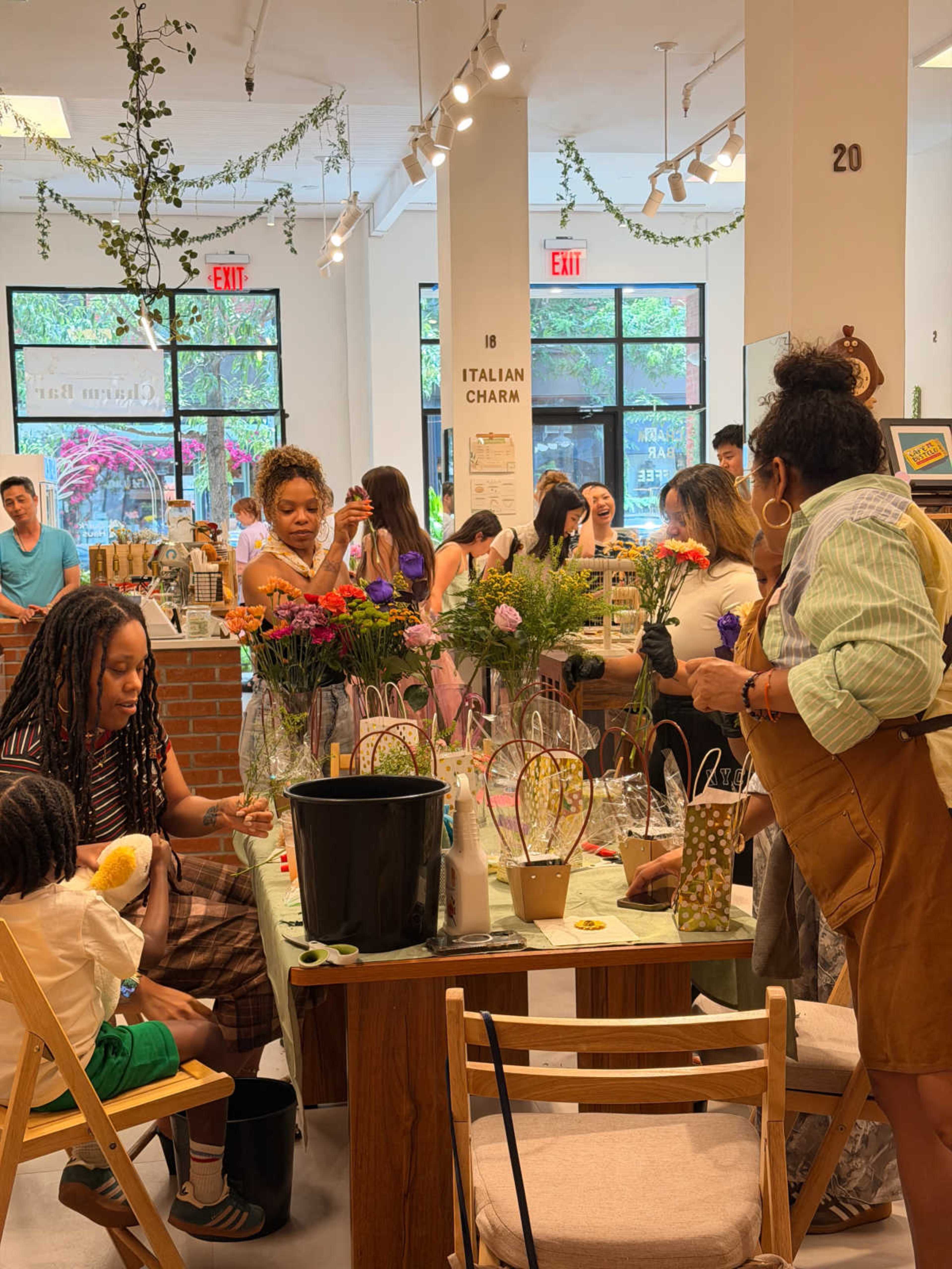 A group of people engages in a floral arrangement workshop inside a well-lit store, surrounded by greenery and colorful decor.