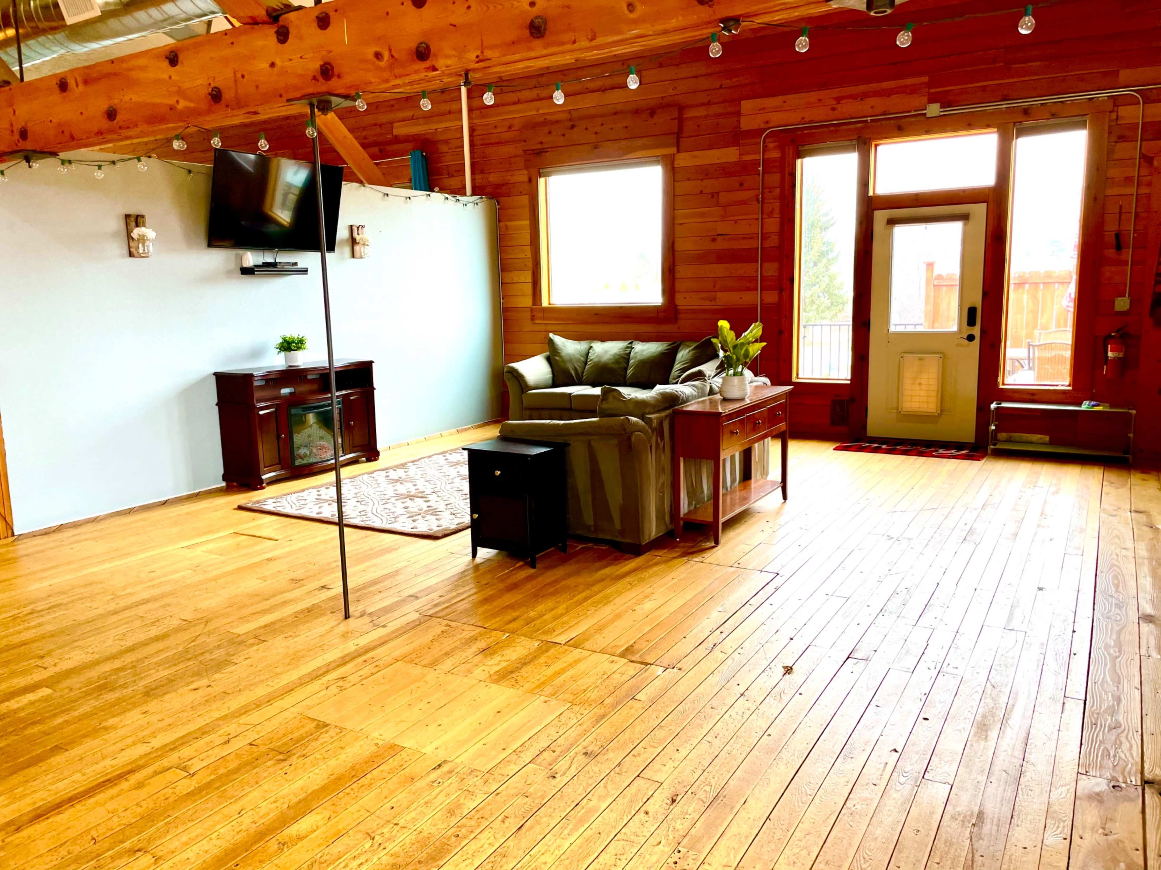 A spacious room with wooden floors, a sofa, a television mounted on the wall, and large windows allowing natural light.