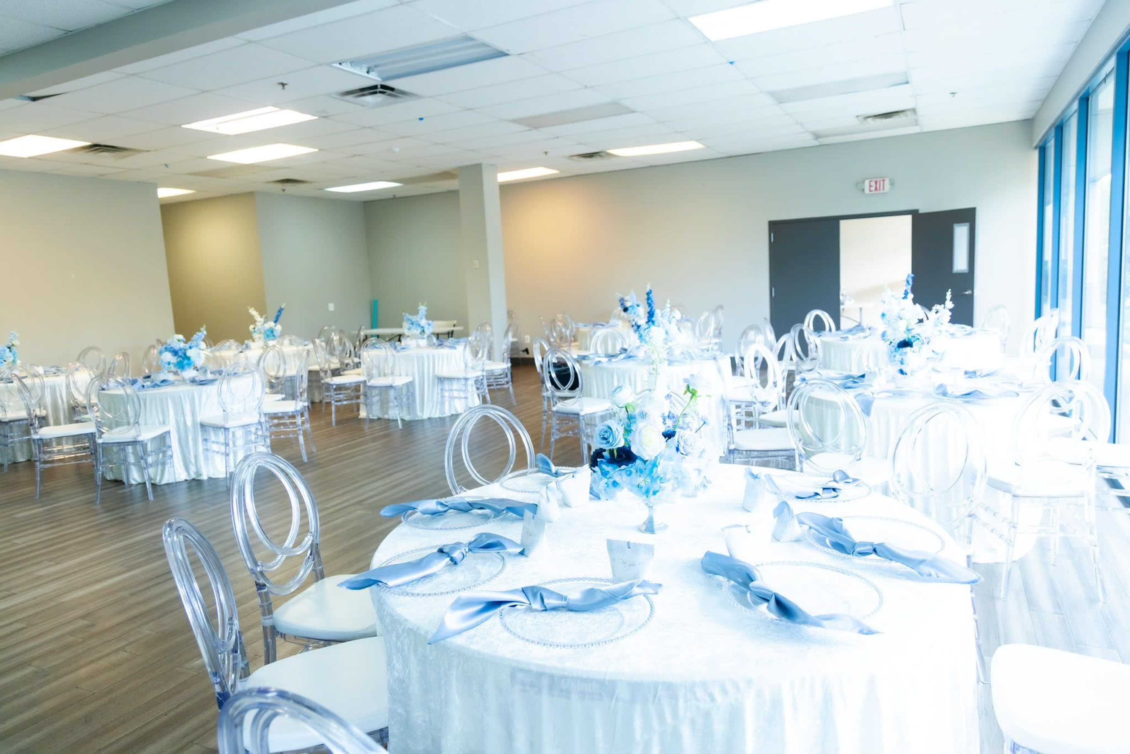 A spacious venue features neatly arranged round tables with white tablecloths and blue decorations, surrounded by clear chairs.