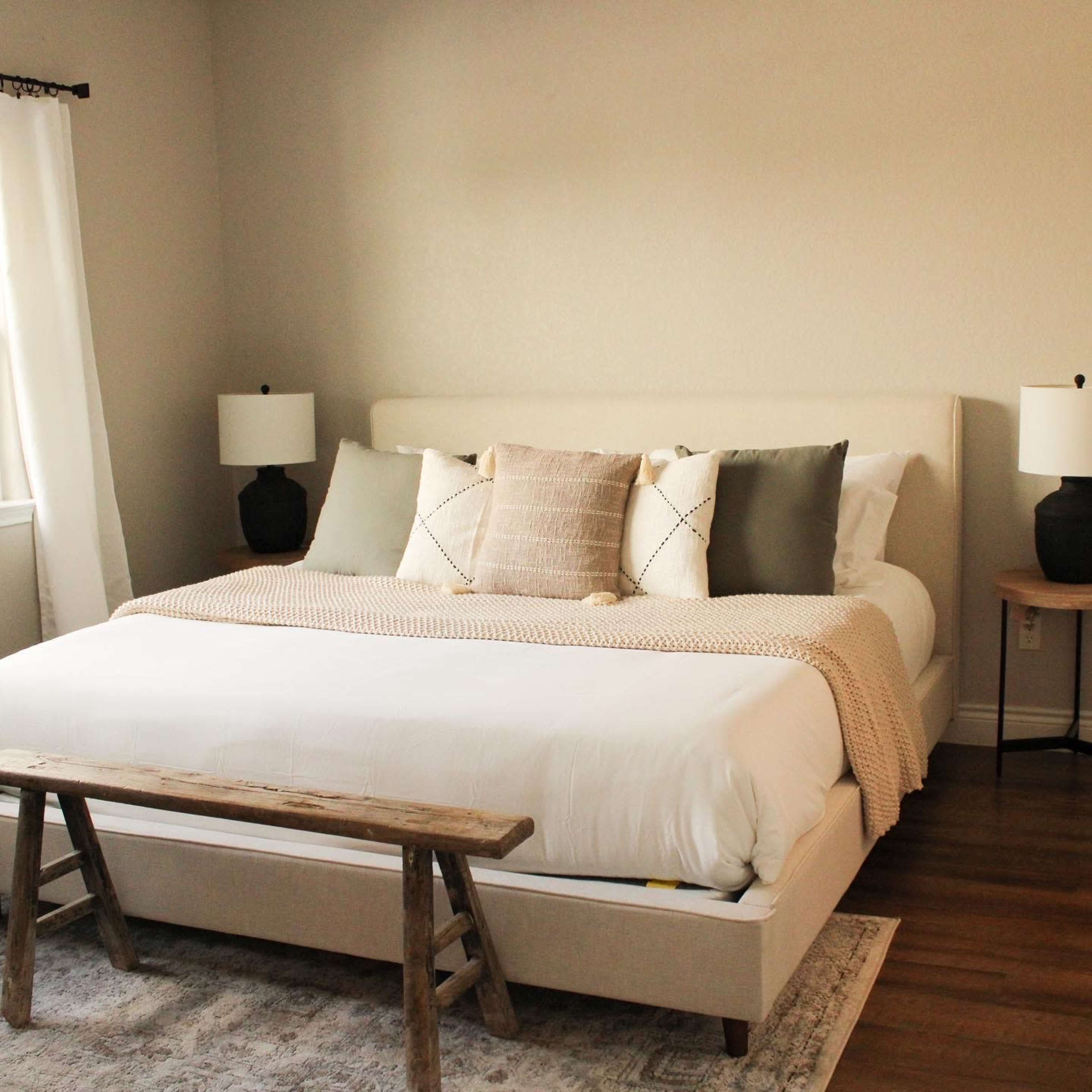 A neatly arranged bedroom with a large bed adorned with pillows and a throw blanket, flanked by two bedside lamps, and a wooden bench at the foot of the bed.