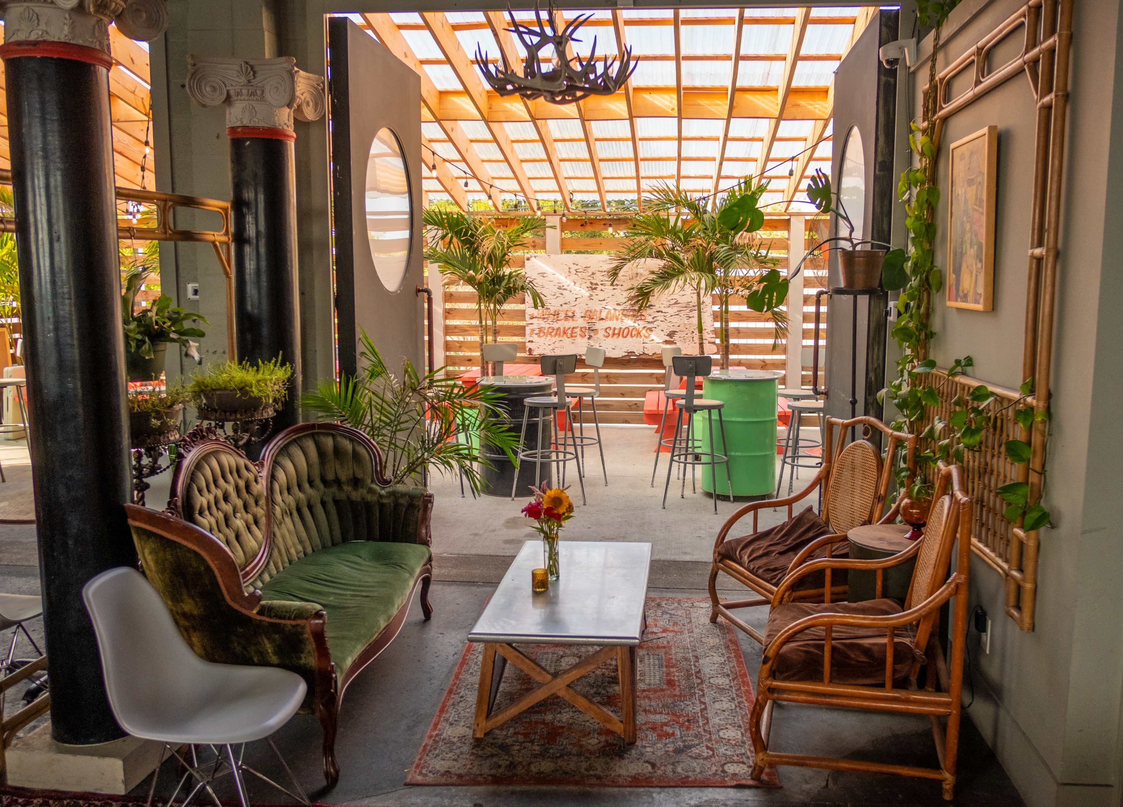 A cozy indoor seating area with vintage furniture, including a green upholstered sofa and cane chairs, leading to a brightly lit outdoor space decorated with plants and tables.