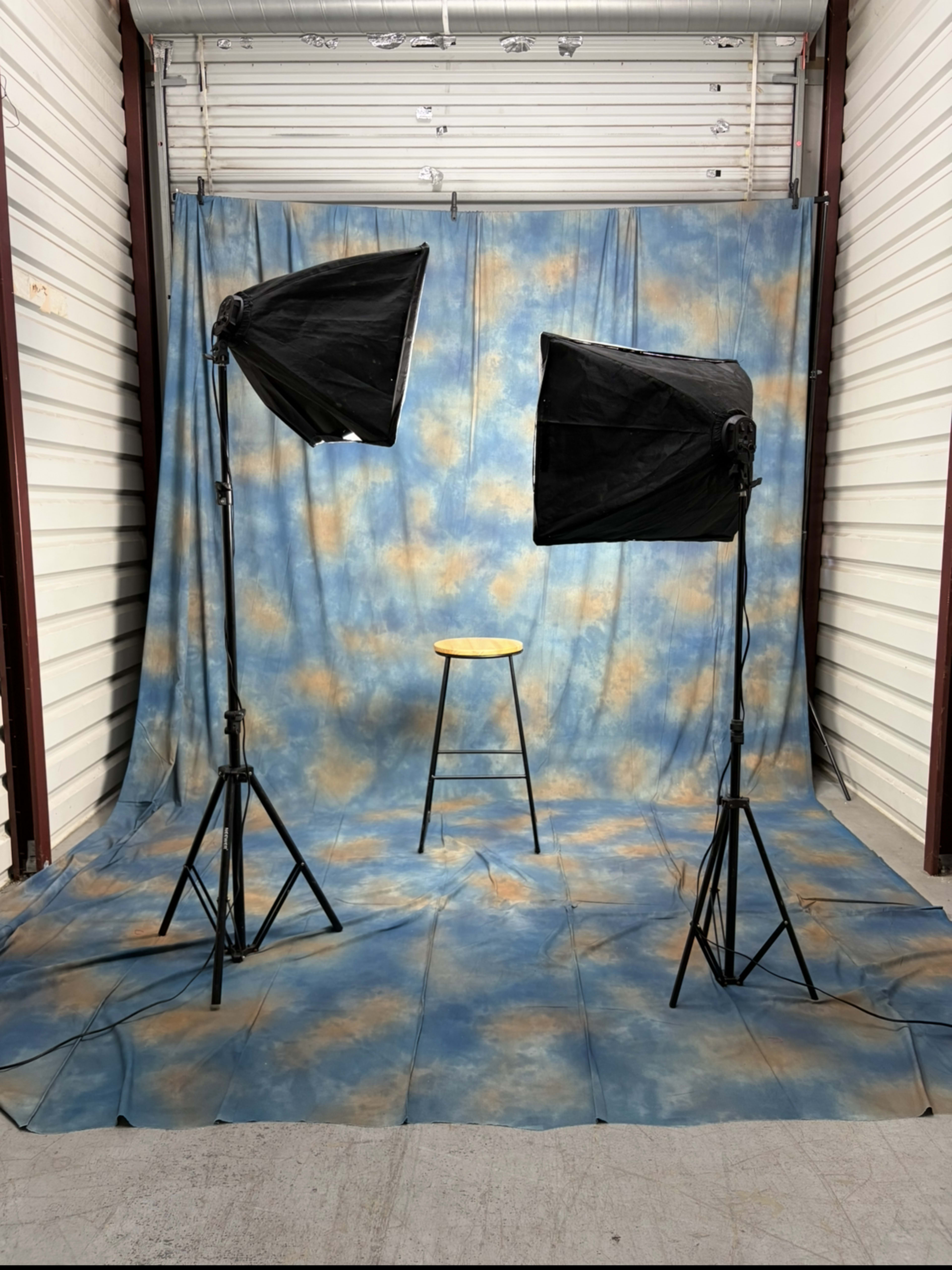 The image shows a photography studio setup featuring a blue and orange backdrop, two light stands with softboxes, and a wooden stool in the center.