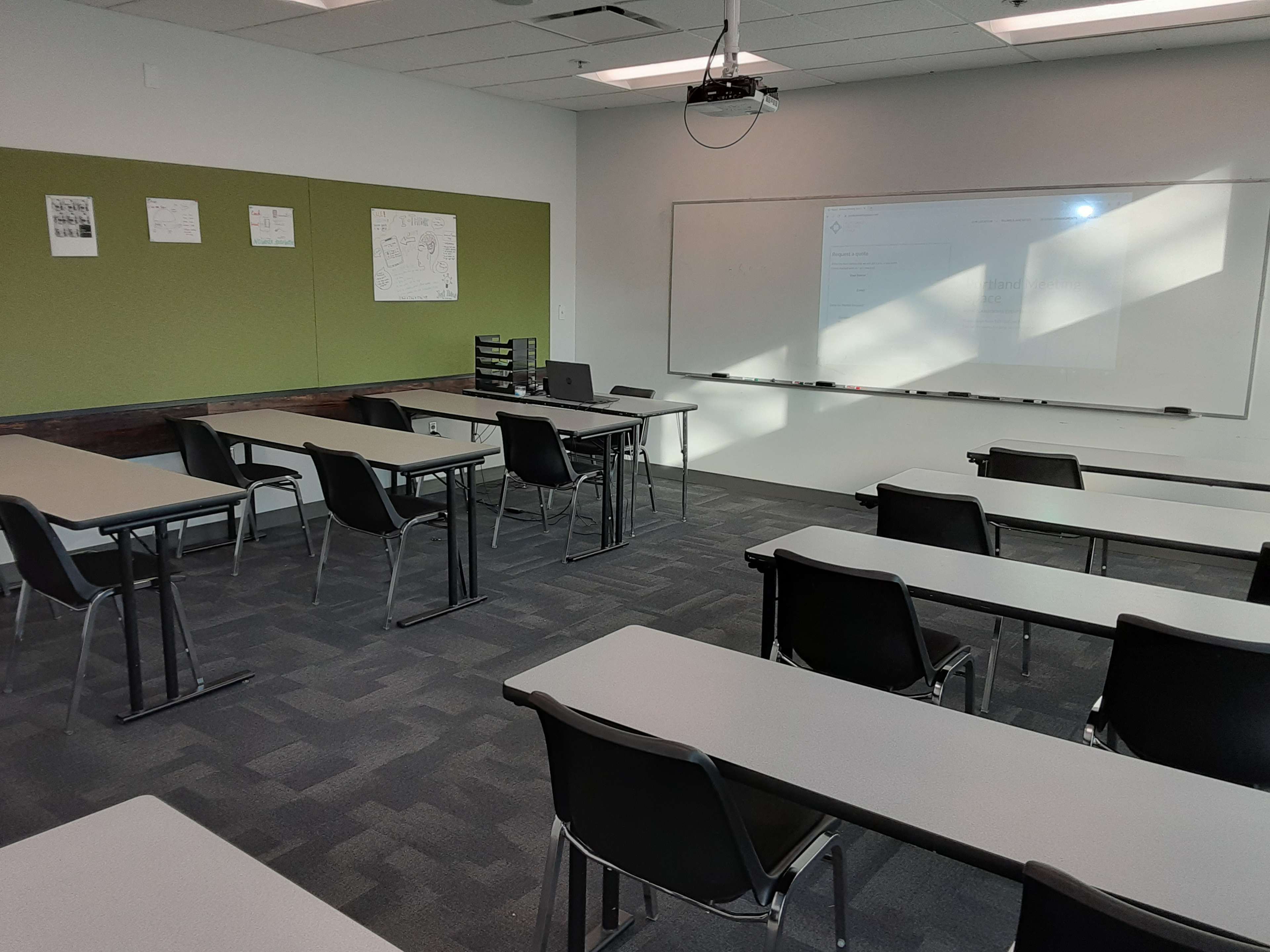 A classroom is arranged with multiple rows of desks facing a whiteboard and projector screen, with some educational materials displayed on the wall.