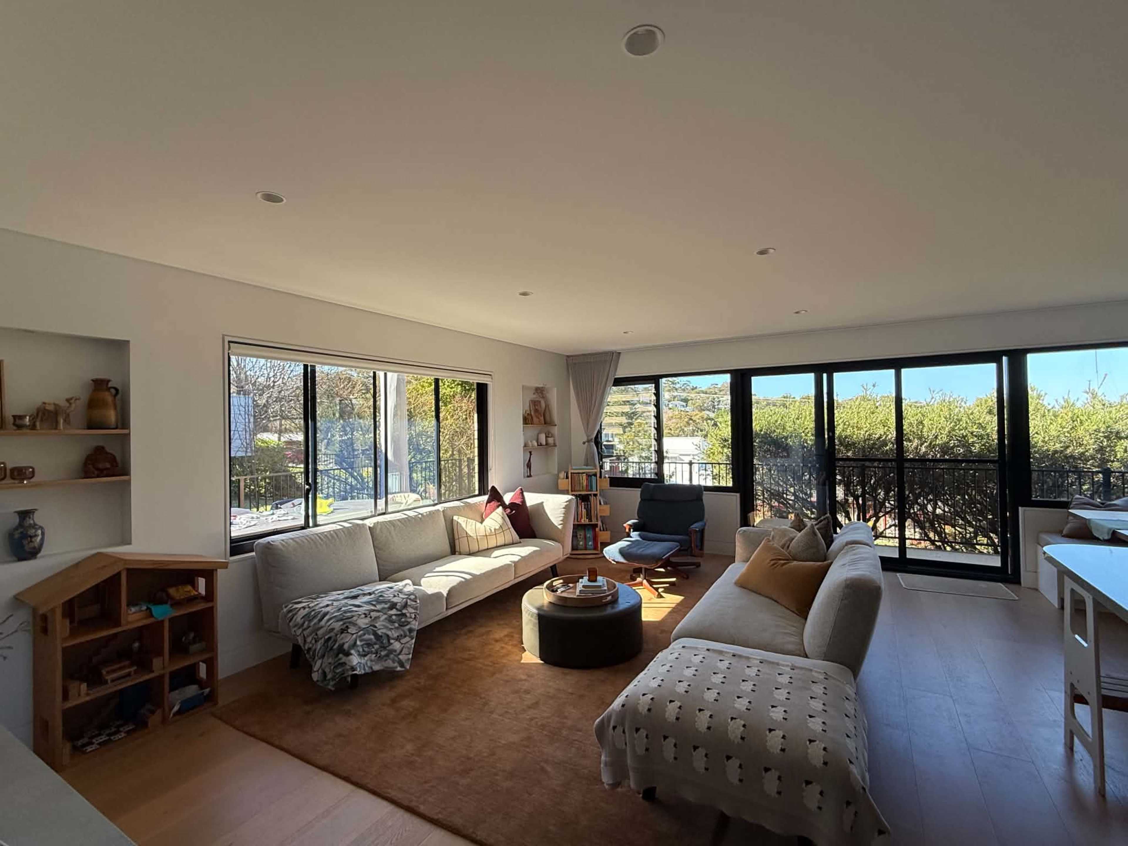 The image shows a modern living room with large windows, two sofas, a wooden table, and a bookshelf.