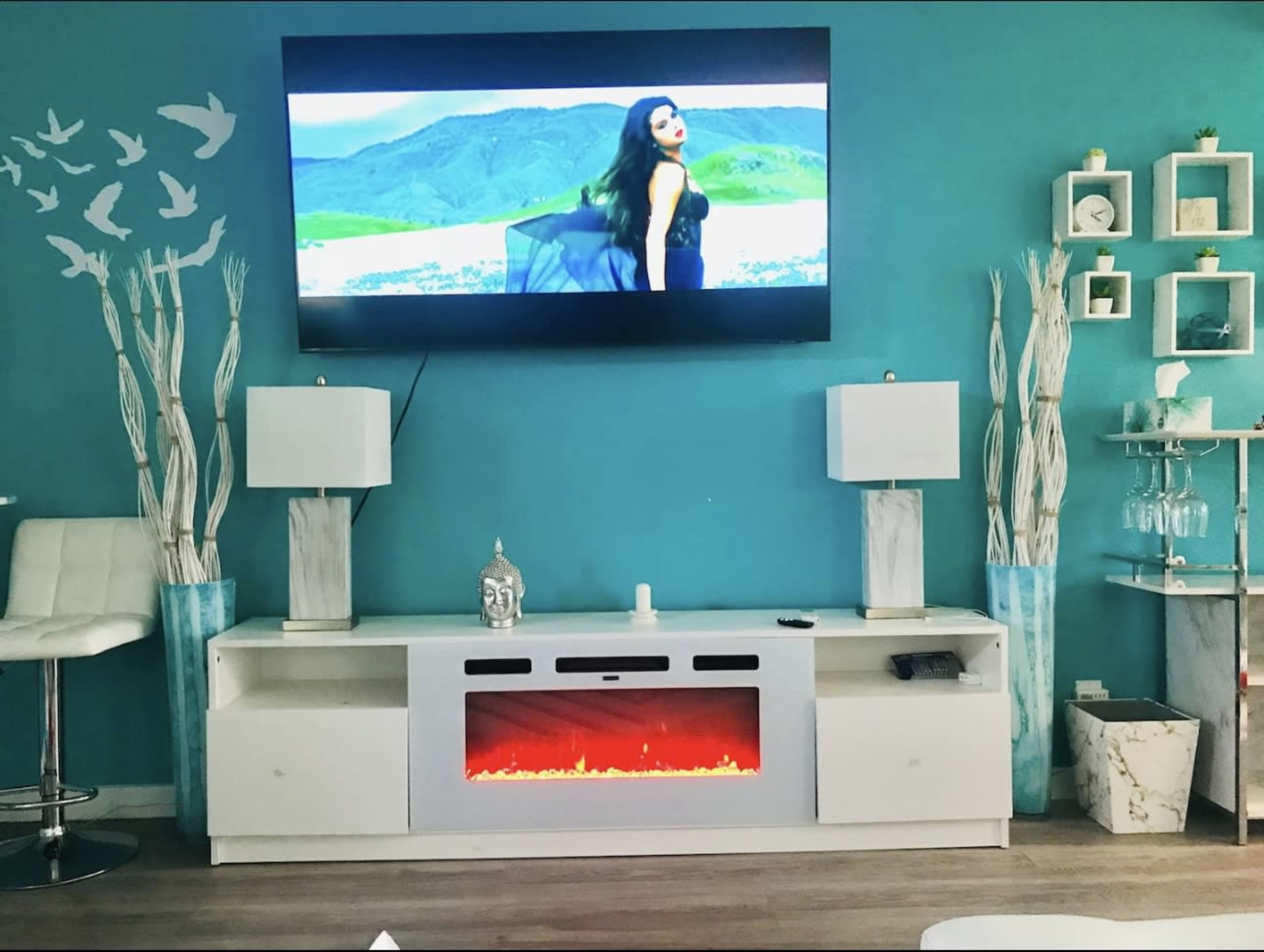 The image shows a modern living room with a turquoise wall, a television mounted above a white entertainment center featuring an electric fireplace, flanked by two white lamps and decorative vases.