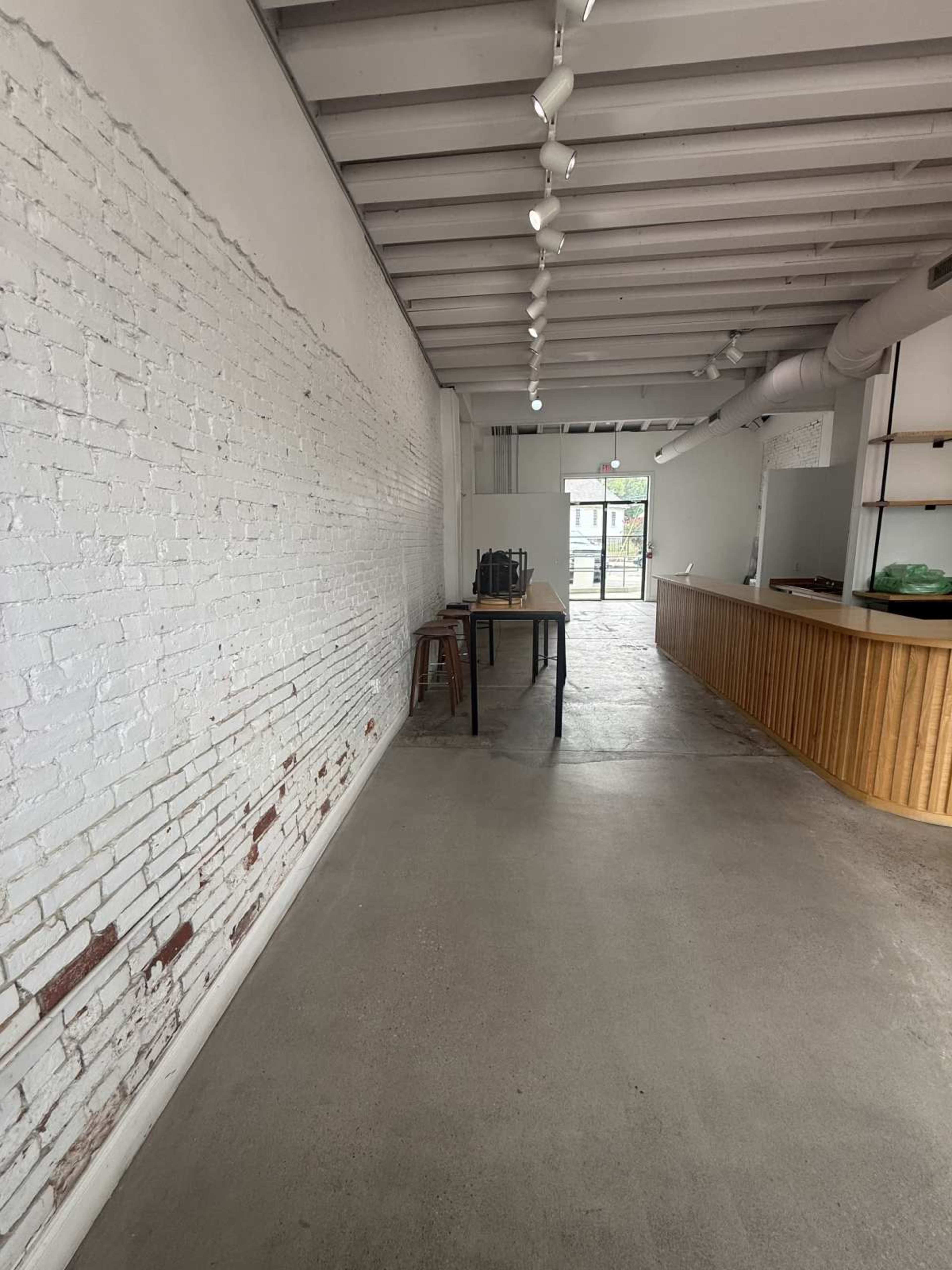 The image shows a spacious interior of a commercial space featuring a white brick wall, concrete floors, and minimal furnishings including a table and stools.