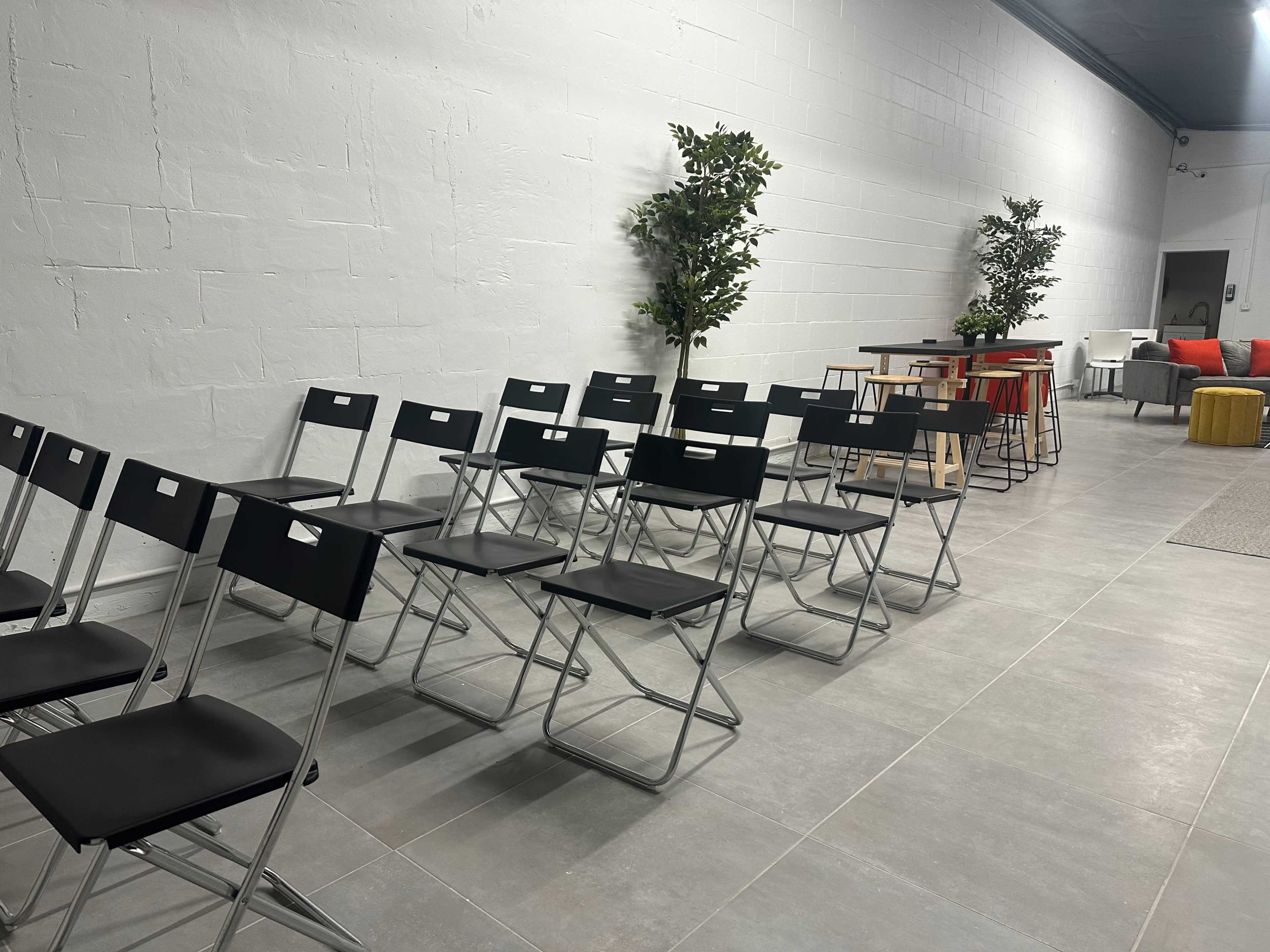 A row of black folding chairs aligned along a wall in a spacious, minimally furnished room with potted plants and various tables in the background.