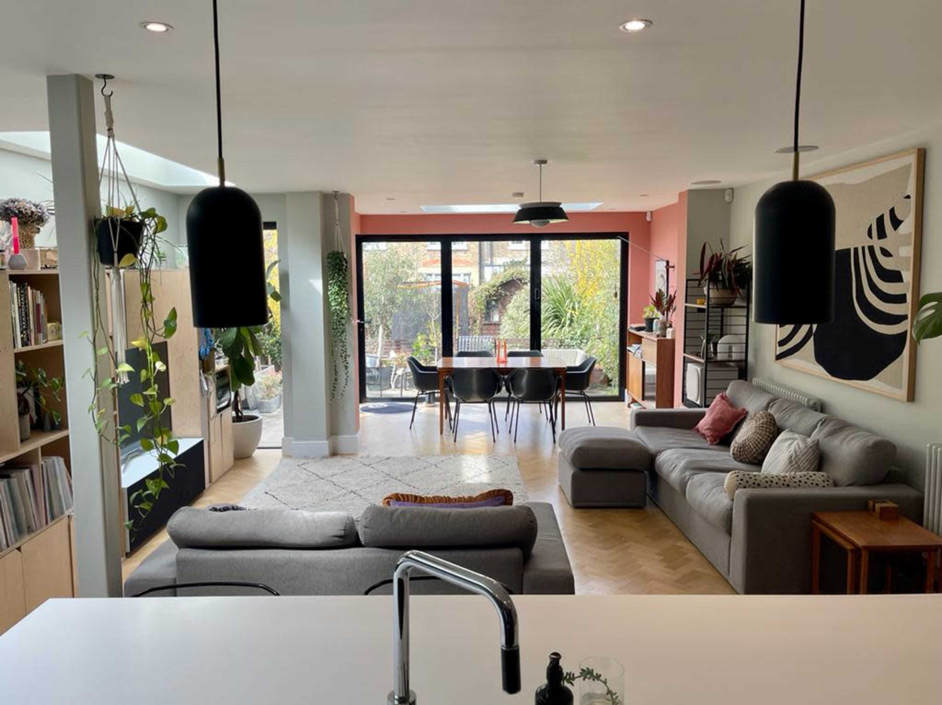 A modern living space features a sectional sofa, a dining area with a black table, and large sliding glass doors opening to a garden, surrounded by plants and contemporary decor.