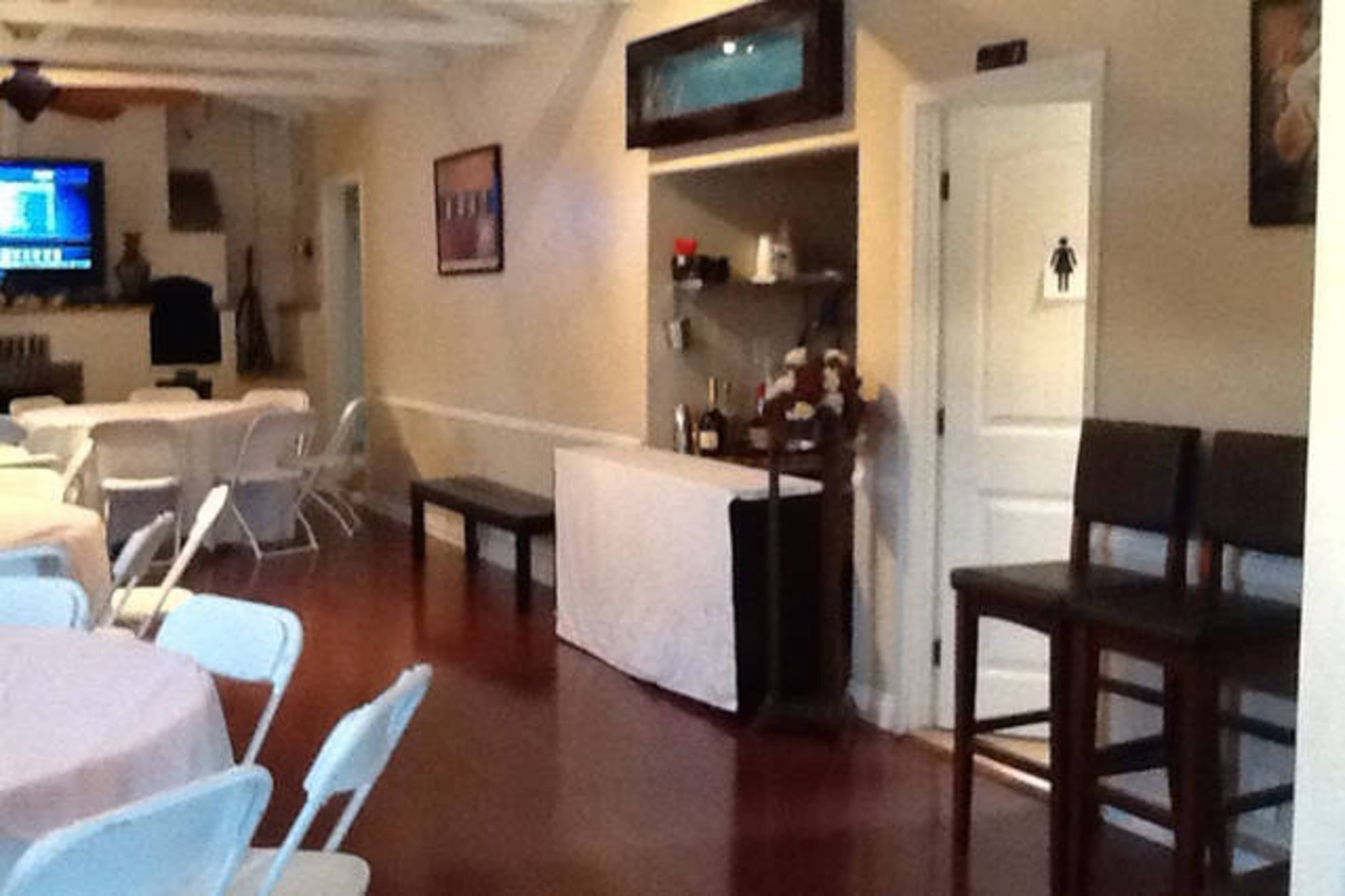 A simple indoor venue with white tables and chairs, a small bar area, and a restroom sign on the wall.