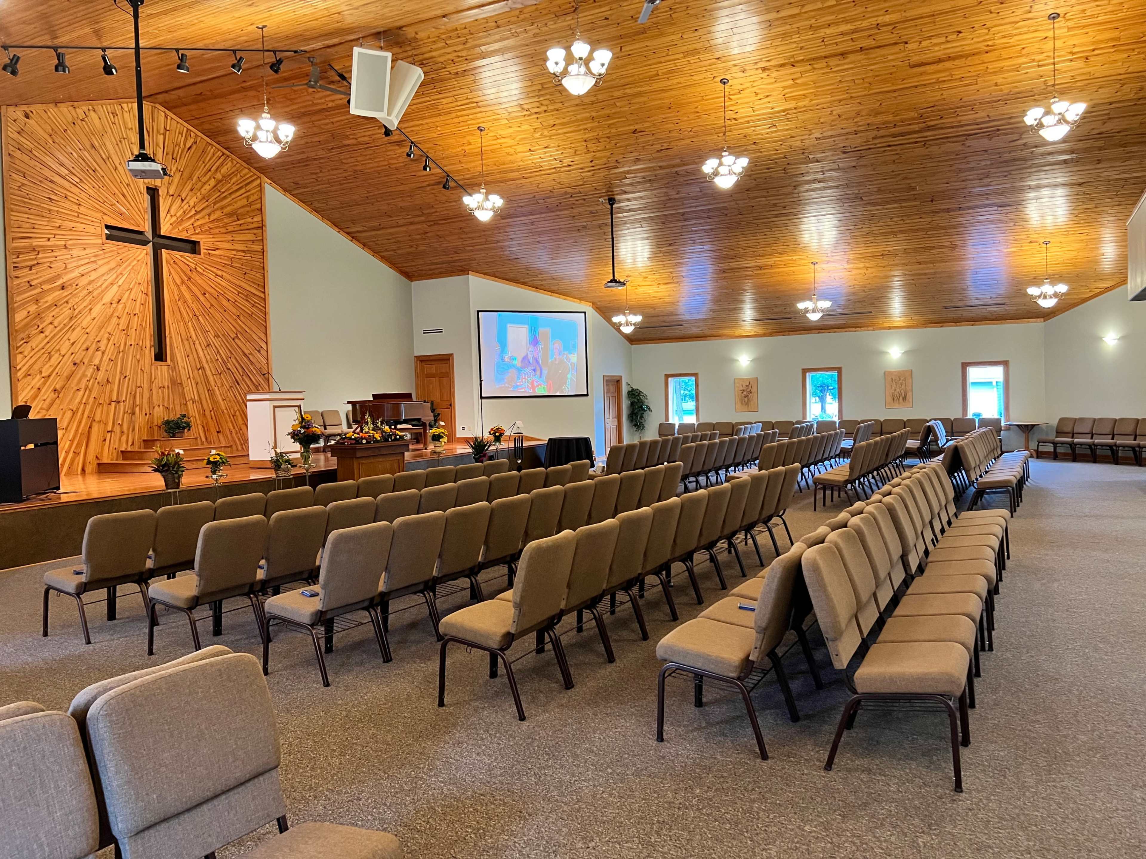 The interior of a church features rows of padded chairs facing a wooden stage with a cross and a projector screen displaying images.