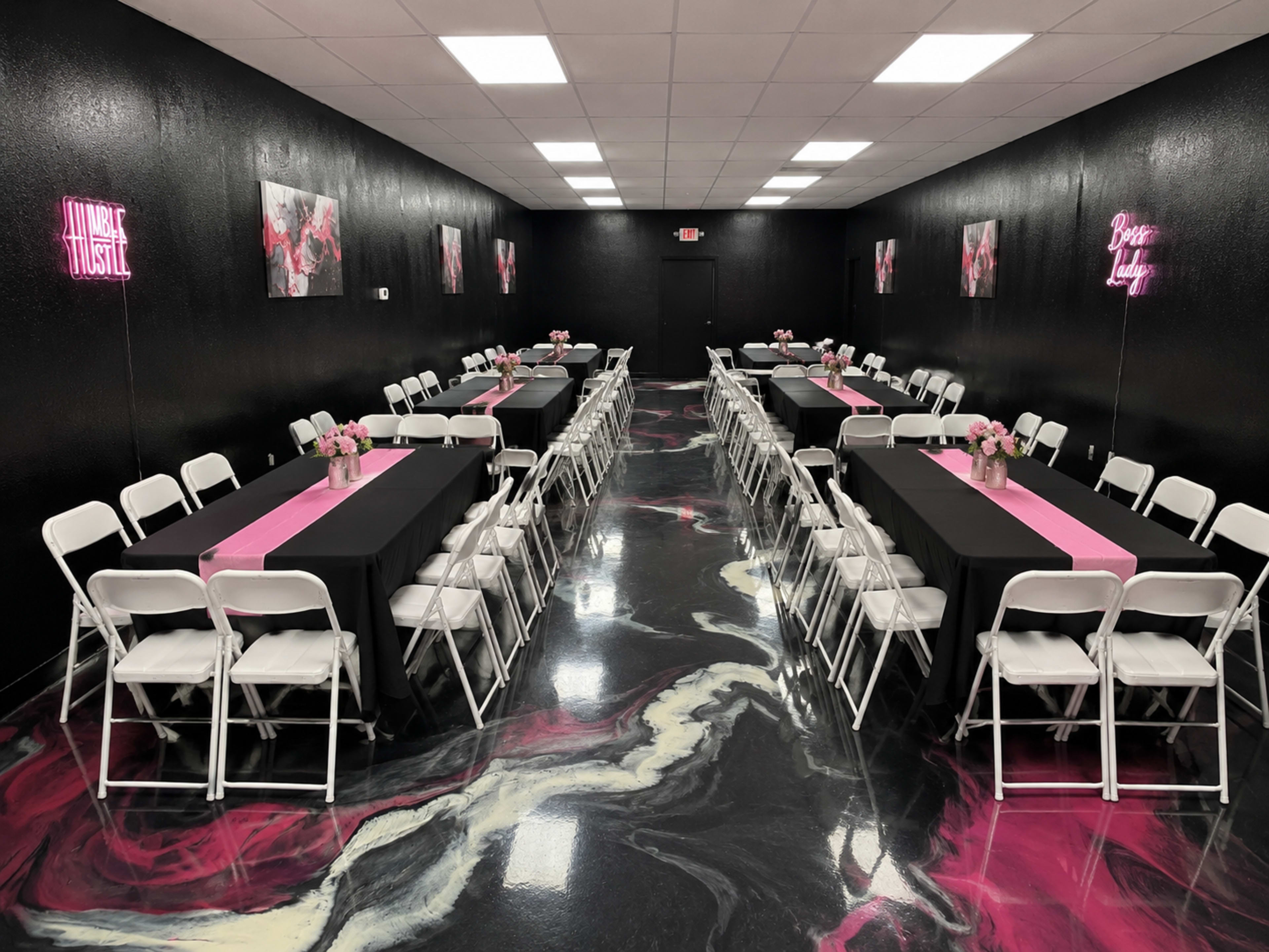 The room features two rows of tables set with black tablecloths and pink runners, surrounded by white chairs, with pink floral centerpieces and neon signs on the walls.