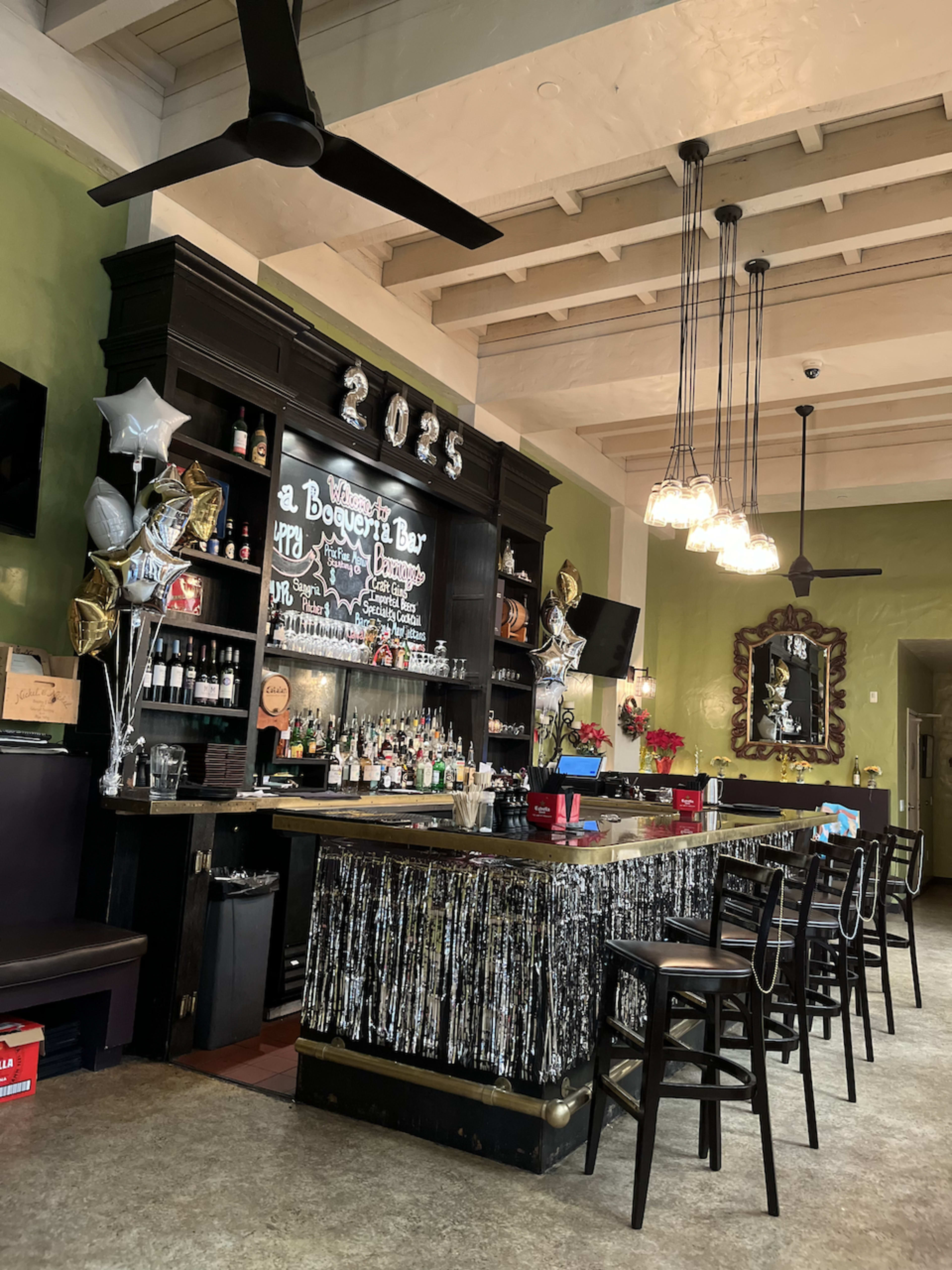The image shows a bar area with a dark wooden counter, shelves of bottles, and festive decorations including balloons and streamers.