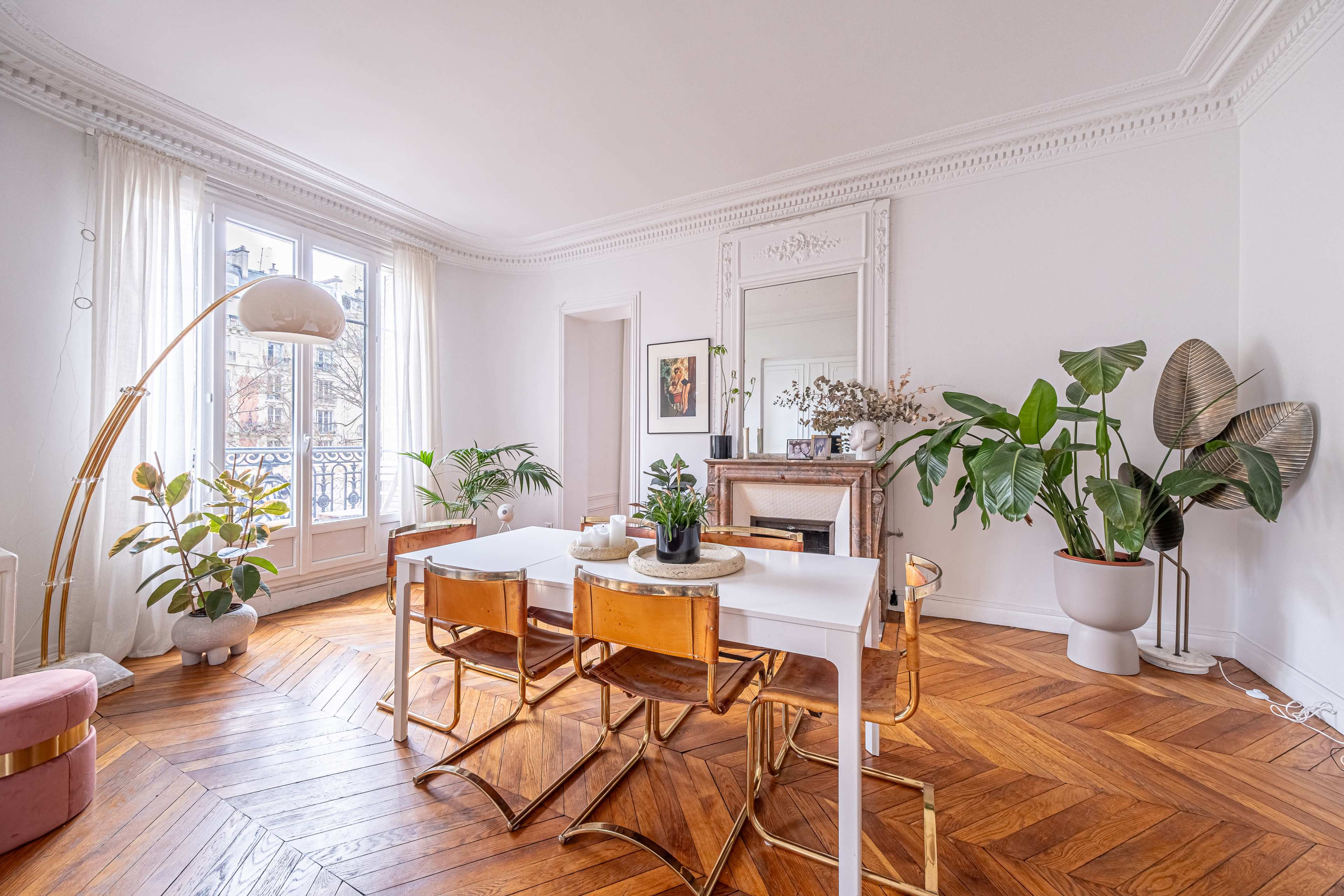 A bright room features a dining table surrounded by four chairs, a large mirror, plants, and hardwood flooring.