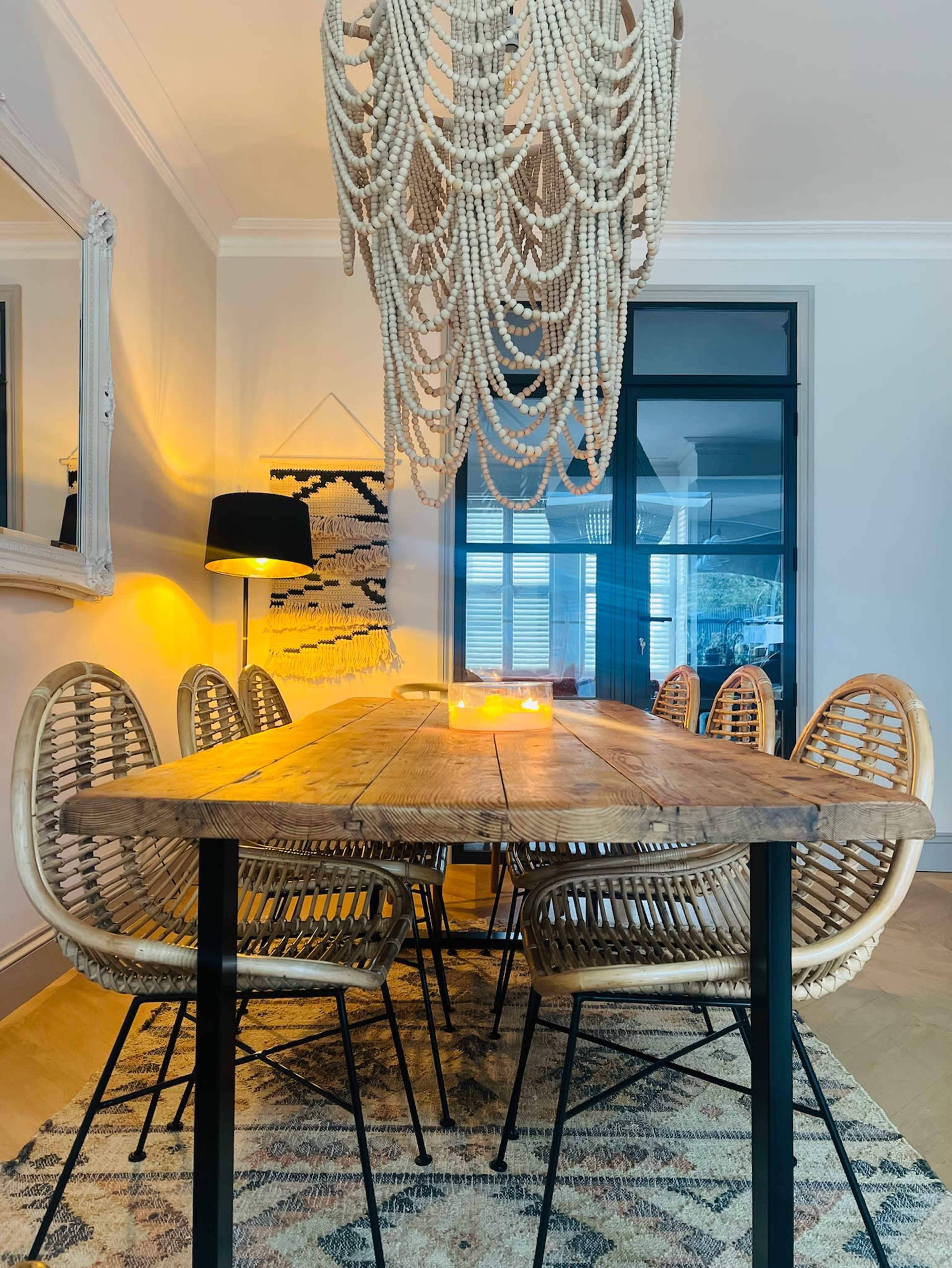 A wooden dining table surrounded by rattan chairs is illuminated by a large beaded chandelier in a softly lit room.