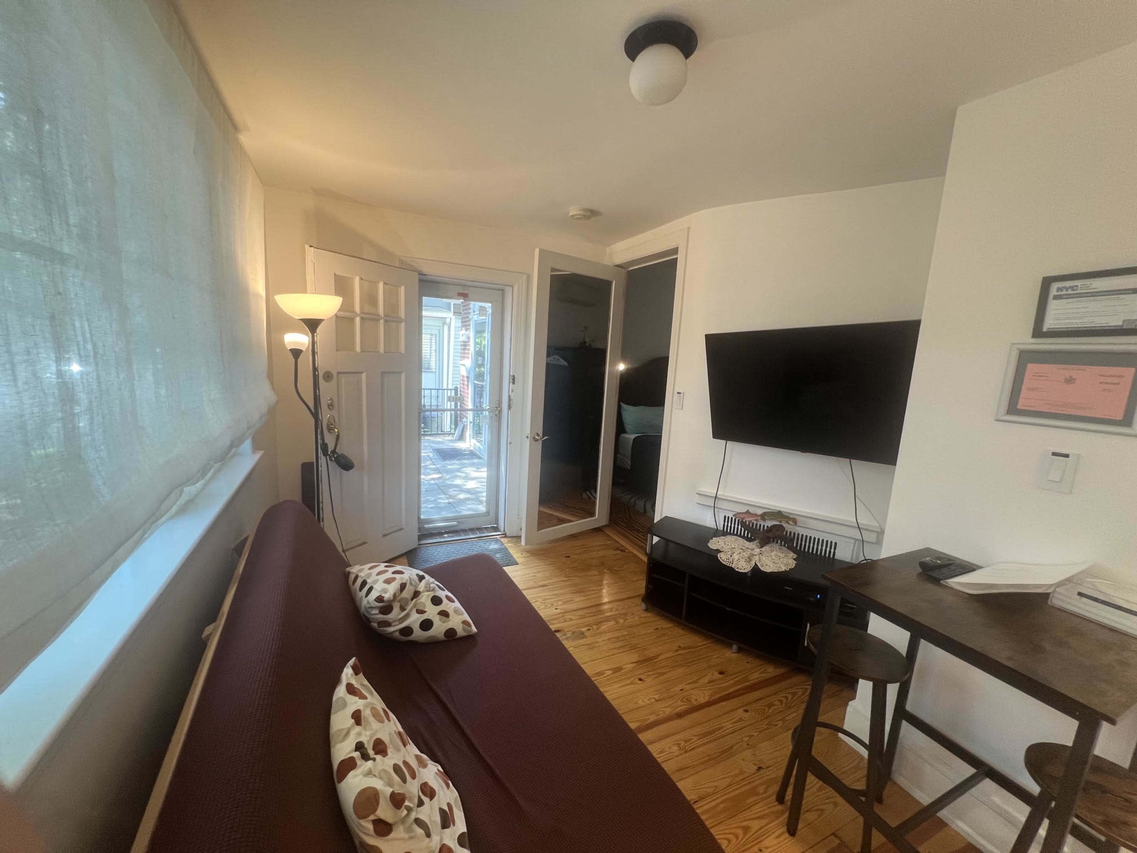 The image shows a small, inviting living space with a brown sofa, a television mounted on the wall, and a view of a doorway leading outdoors.
