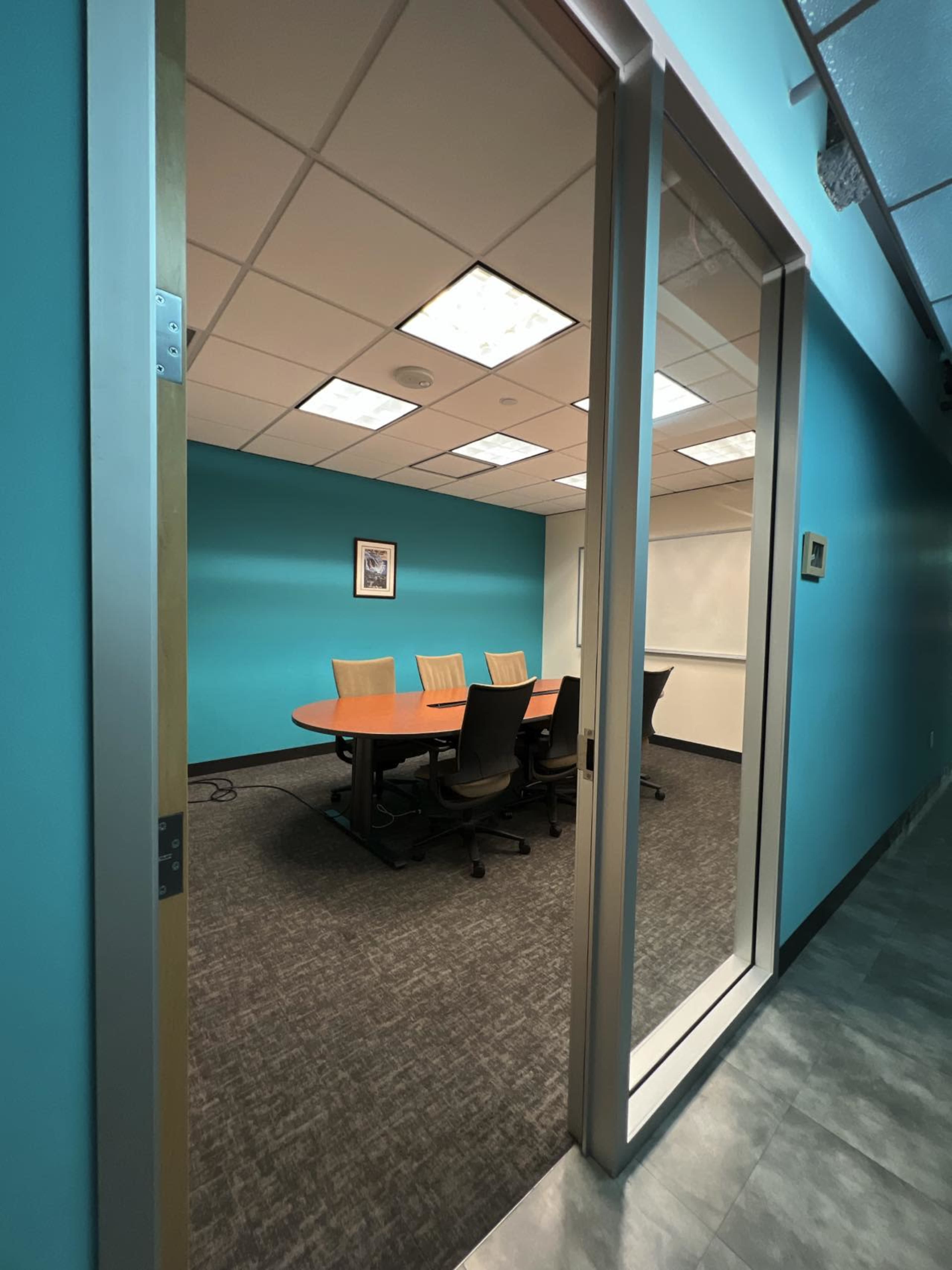 A conference room with a circular table and chairs is visible through a glass door, surrounded by turquoise walls and bright lighting.