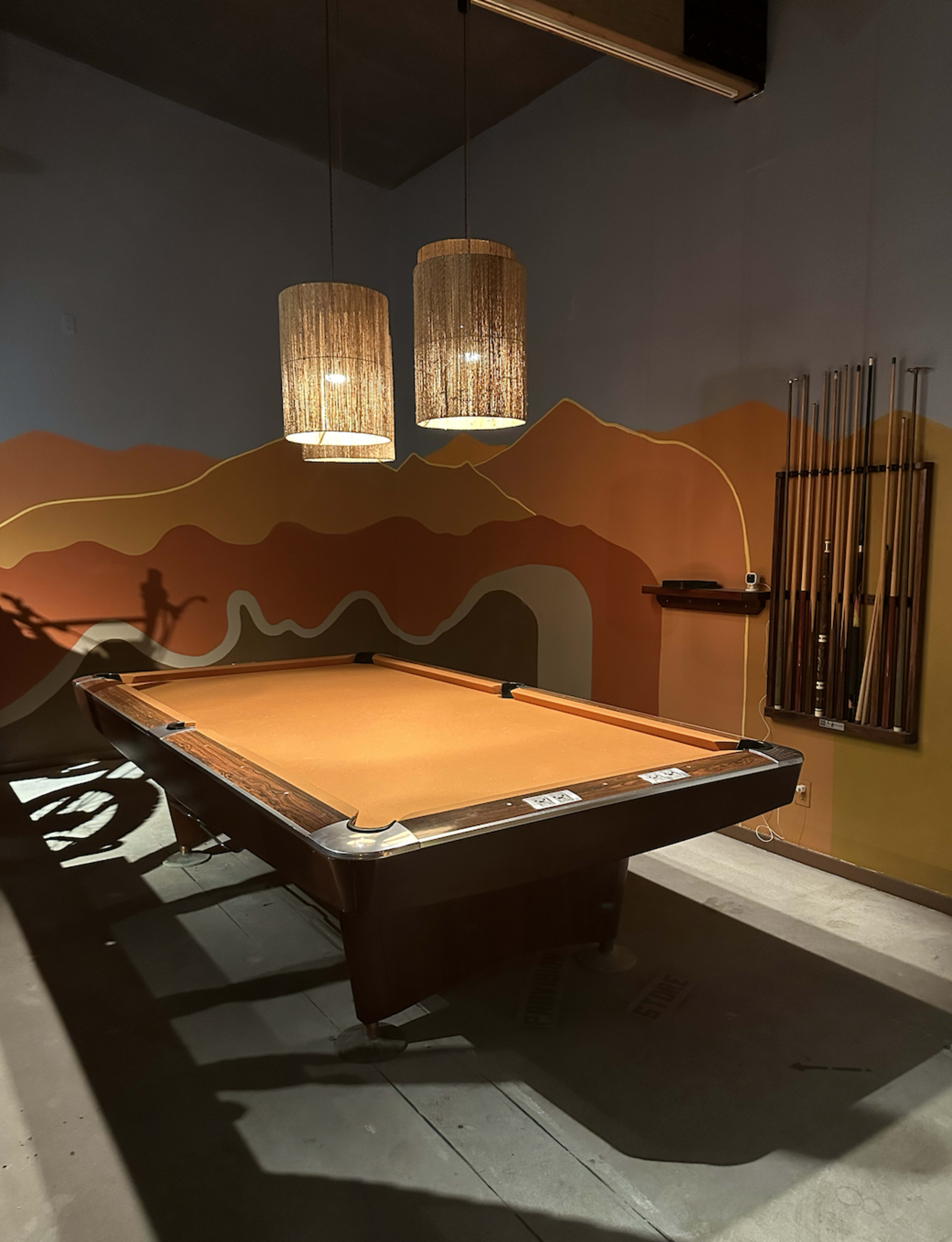 The image shows a pool table set in a dimly lit room with two hanging light fixtures and a colorful mural on the wall.