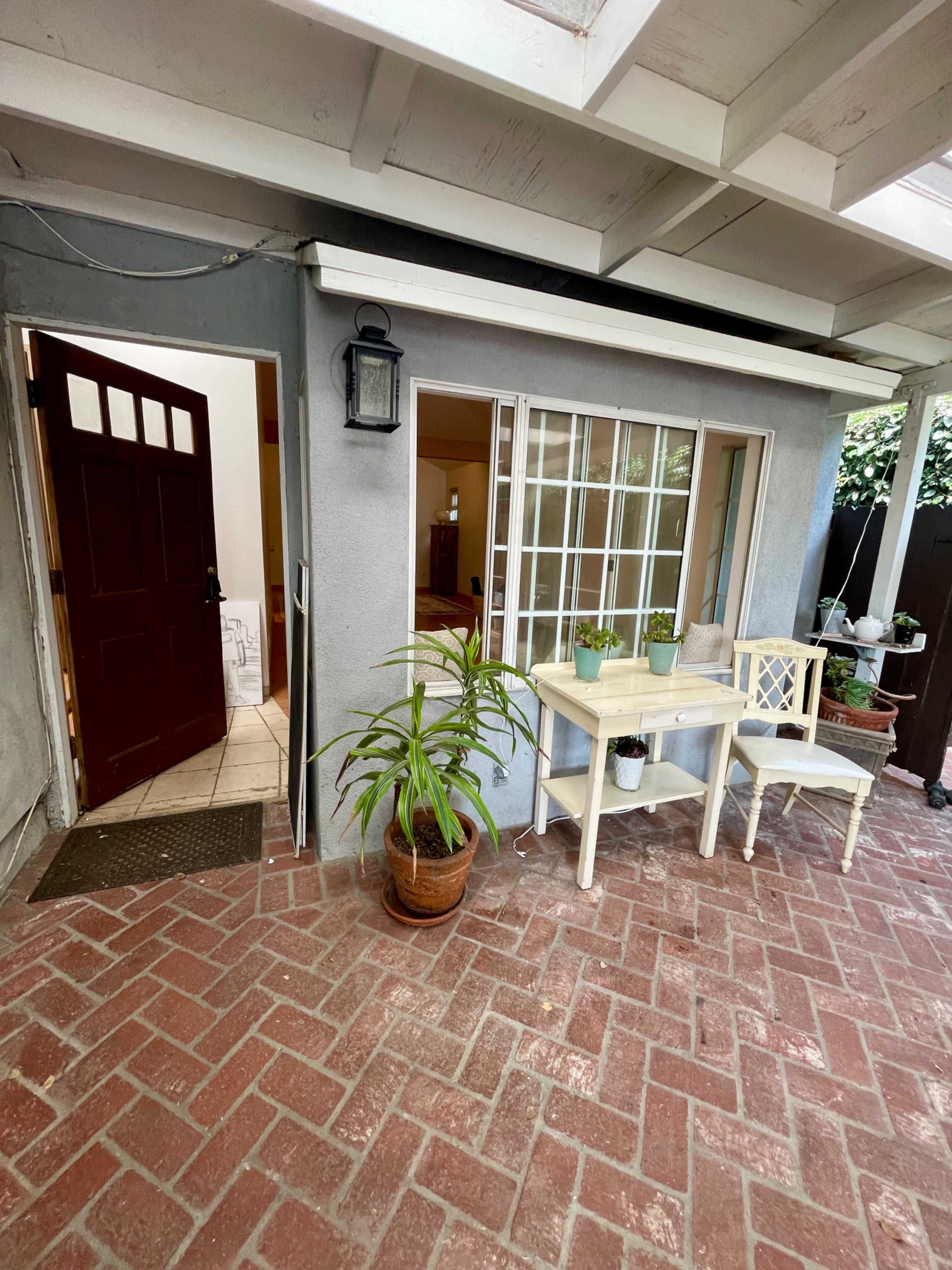 The image shows a small outdoor seating area with a table and chairs next to an entrance door, surrounded by potted plants and a brick floor.
