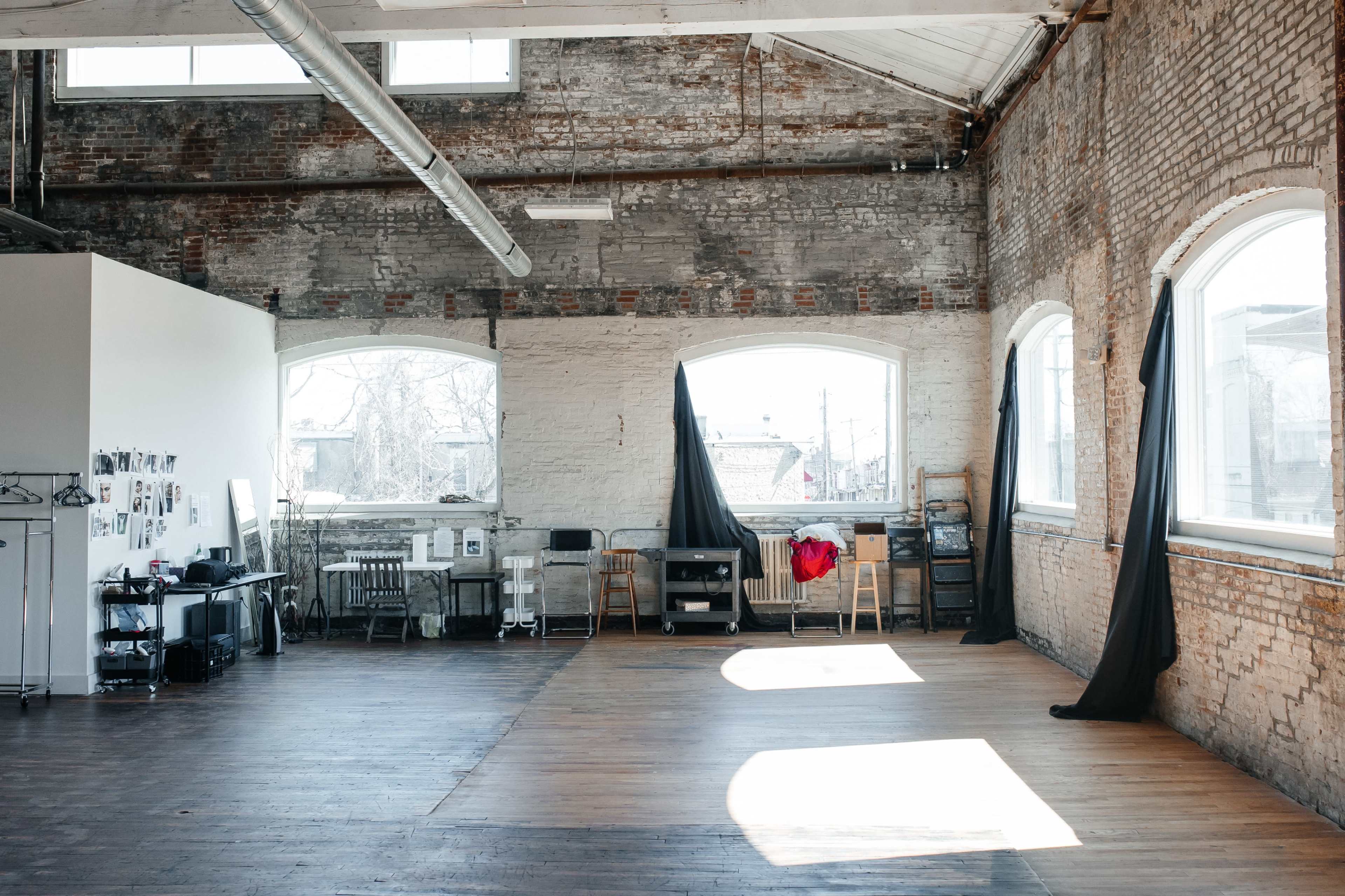 A spacious, industrial-style room with exposed brick walls, large windows, and wooden flooring, featuring some furniture and draped fabric.