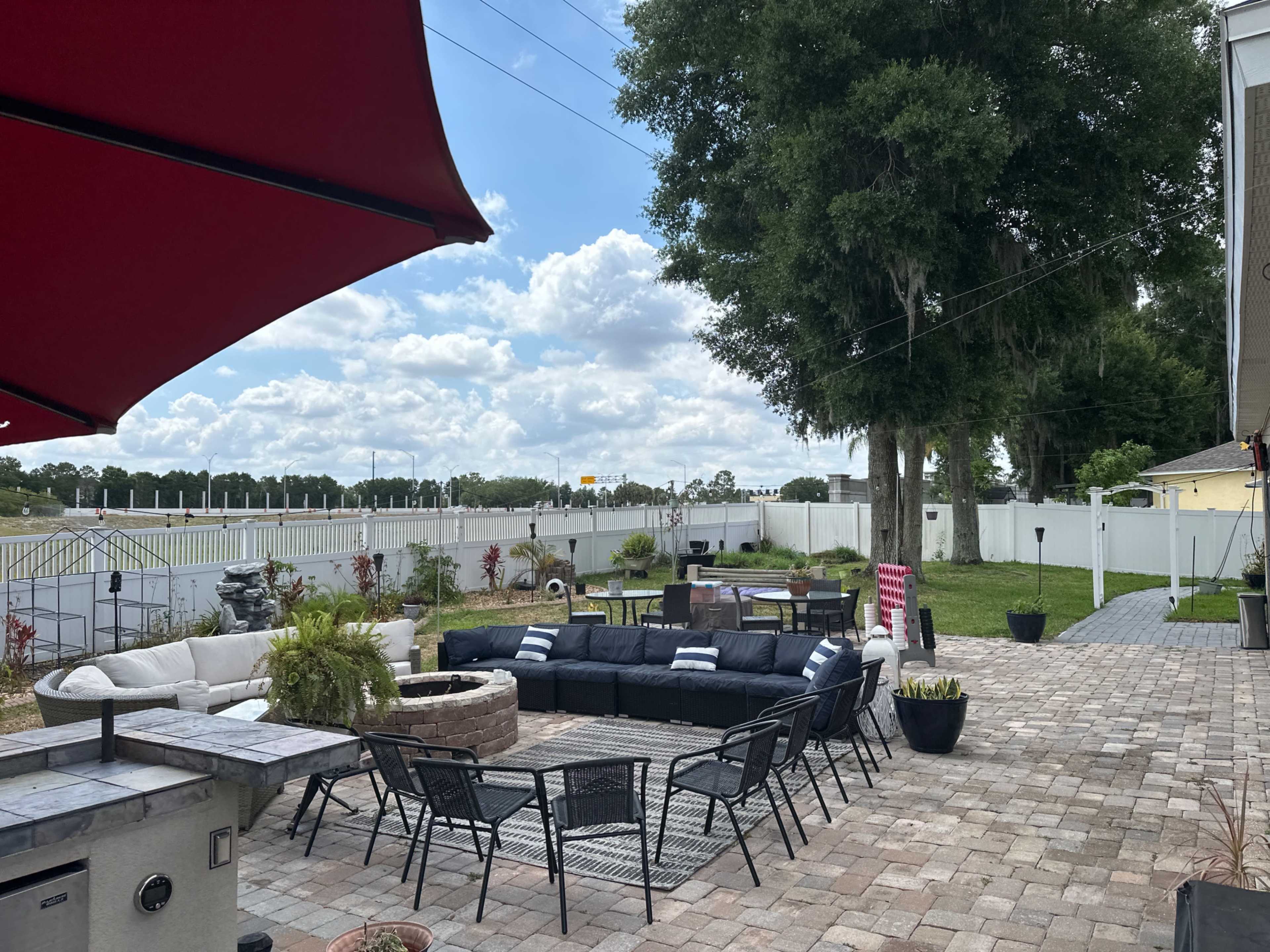 A backyard patio features multiple seating areas with lounge chairs, a sectional sofa, and potted plants, bordered by a fence and dotted with trees under a partly cloudy sky.