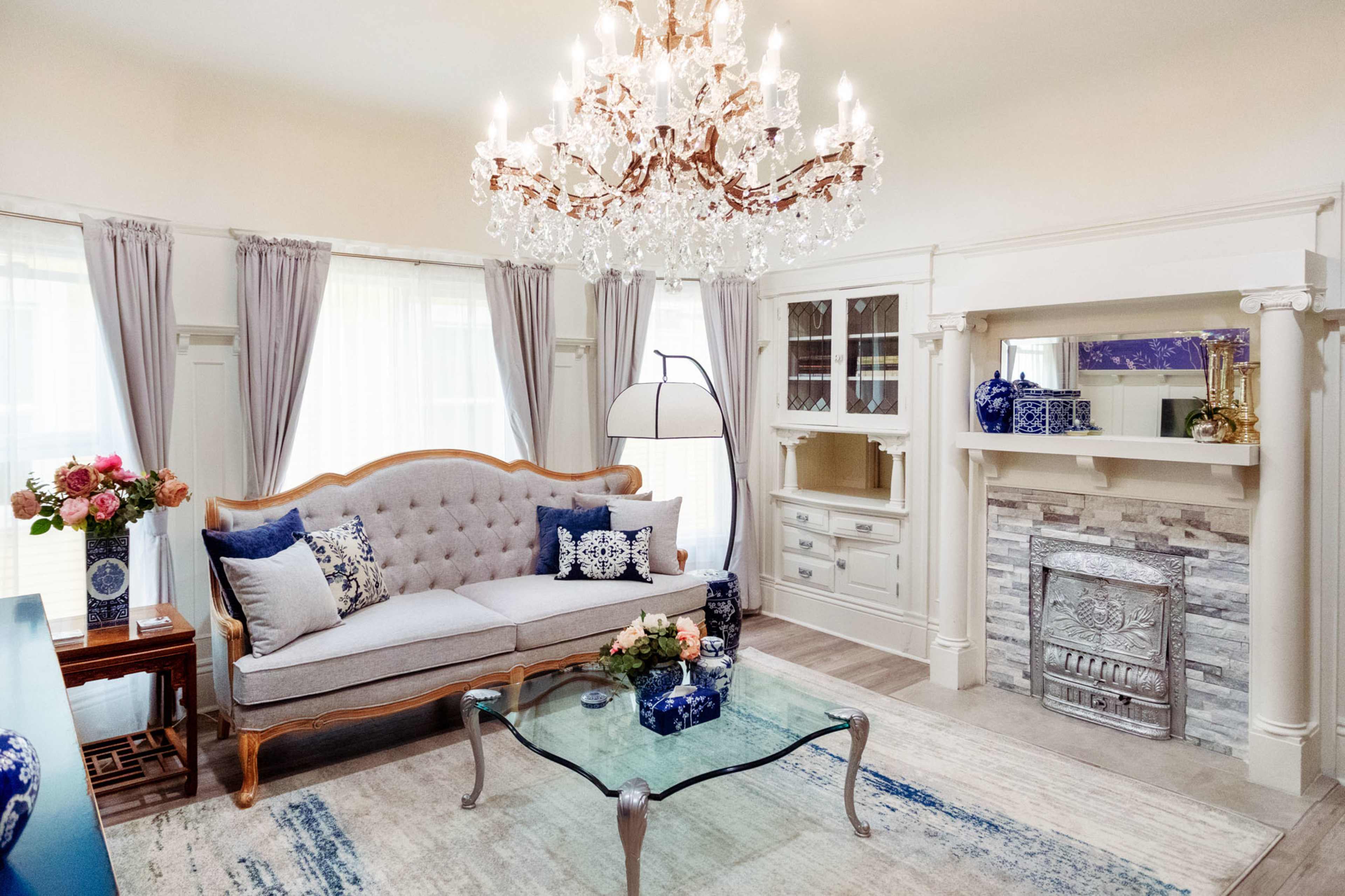The living room features a light gray tufted sofa, a glass coffee table, and a chandelier above, with decorative vases and floral arrangements accenting the decor.