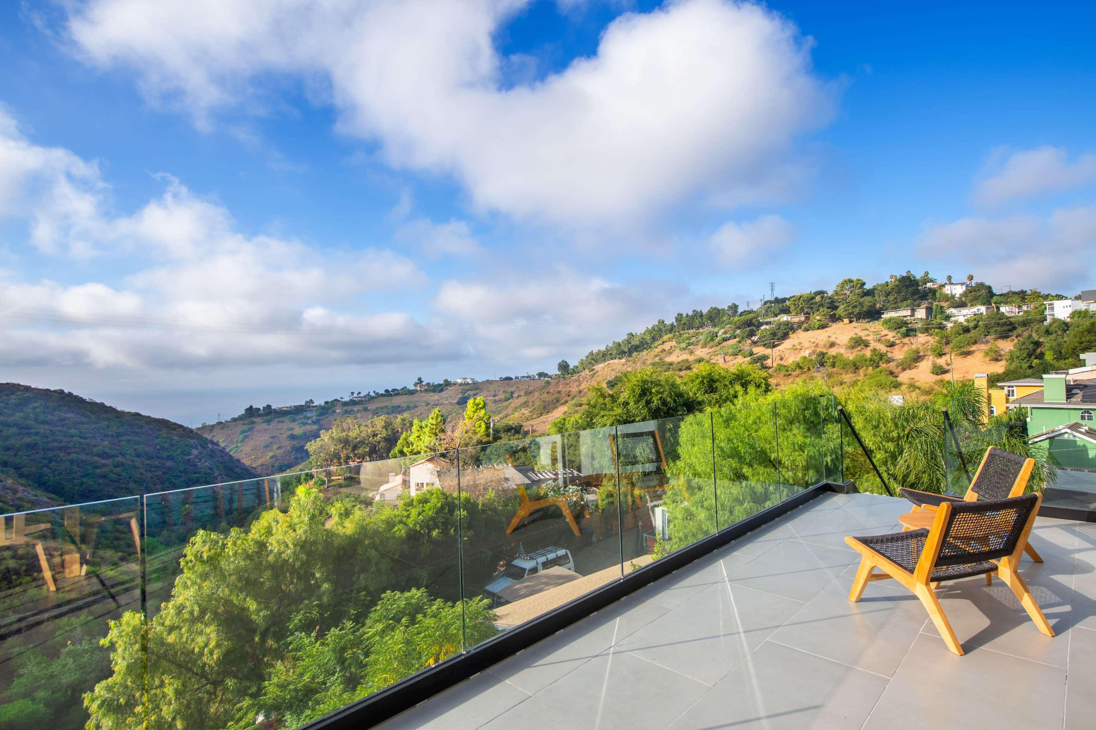A clear view of a hilly landscape is visible from a balcony featuring a wooden chair and glass railing.