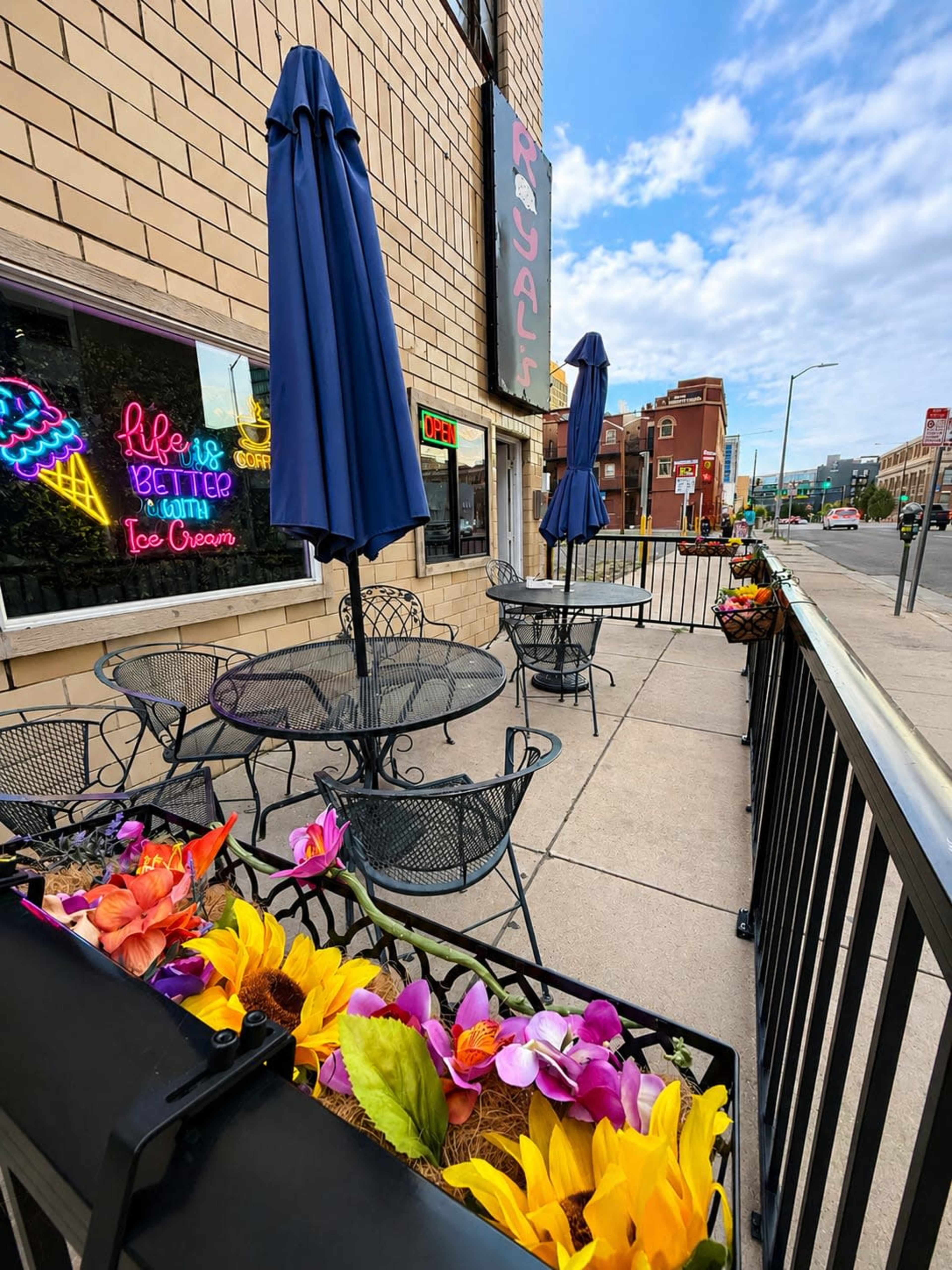 The image shows an outdoor seating area outside a restaurant, featuring black metal tables and chairs along with large blue umbrellas, and colorful flower arrangements along the railing.