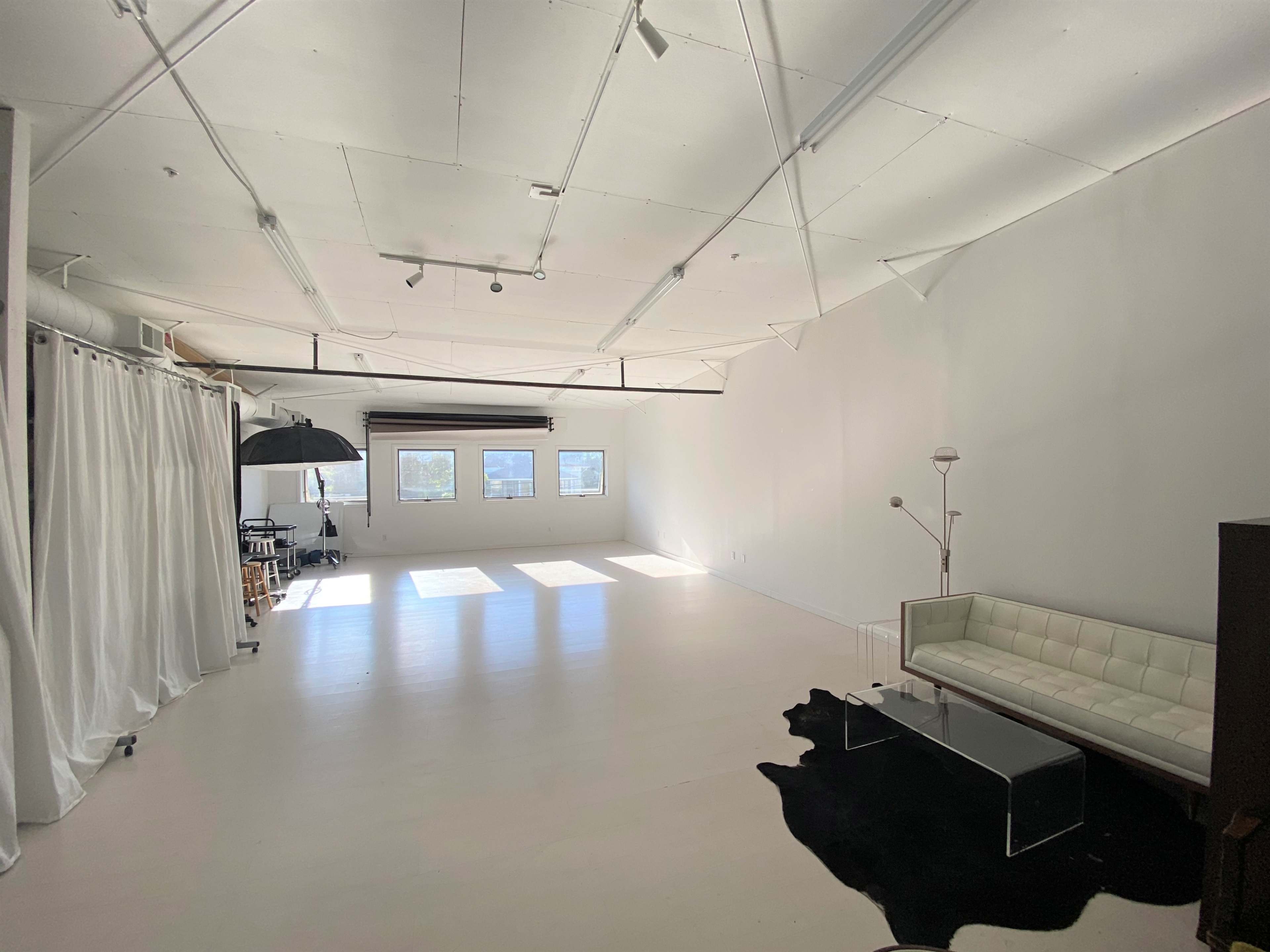 A spacious, minimalist studio with white walls, a large window, a black and white rug, a white sofa, and a modern floor lamp.