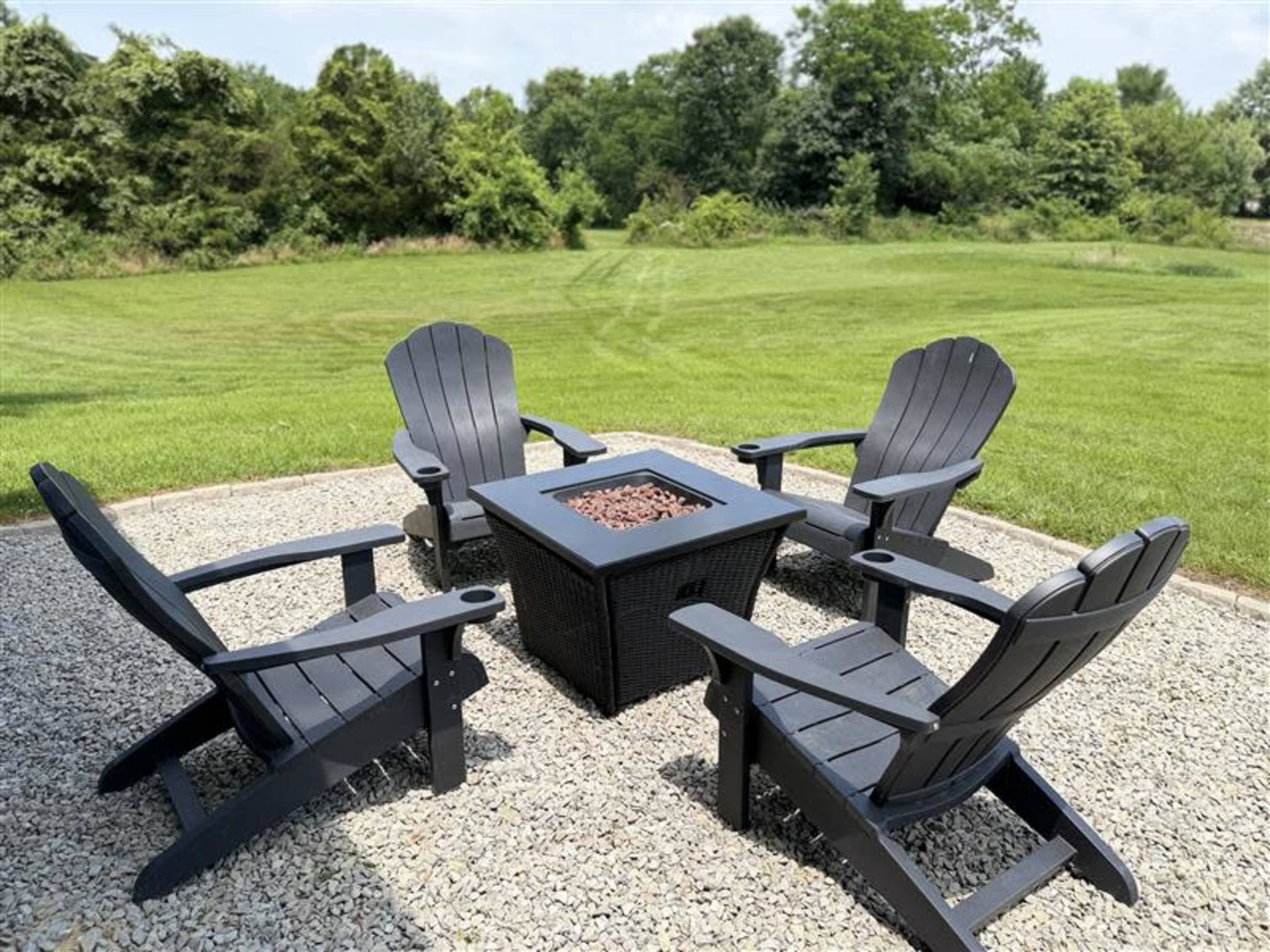 A set of four black Adirondack chairs surrounds a square fire pit on a gravel patio, with a lush green lawn and trees in the background.