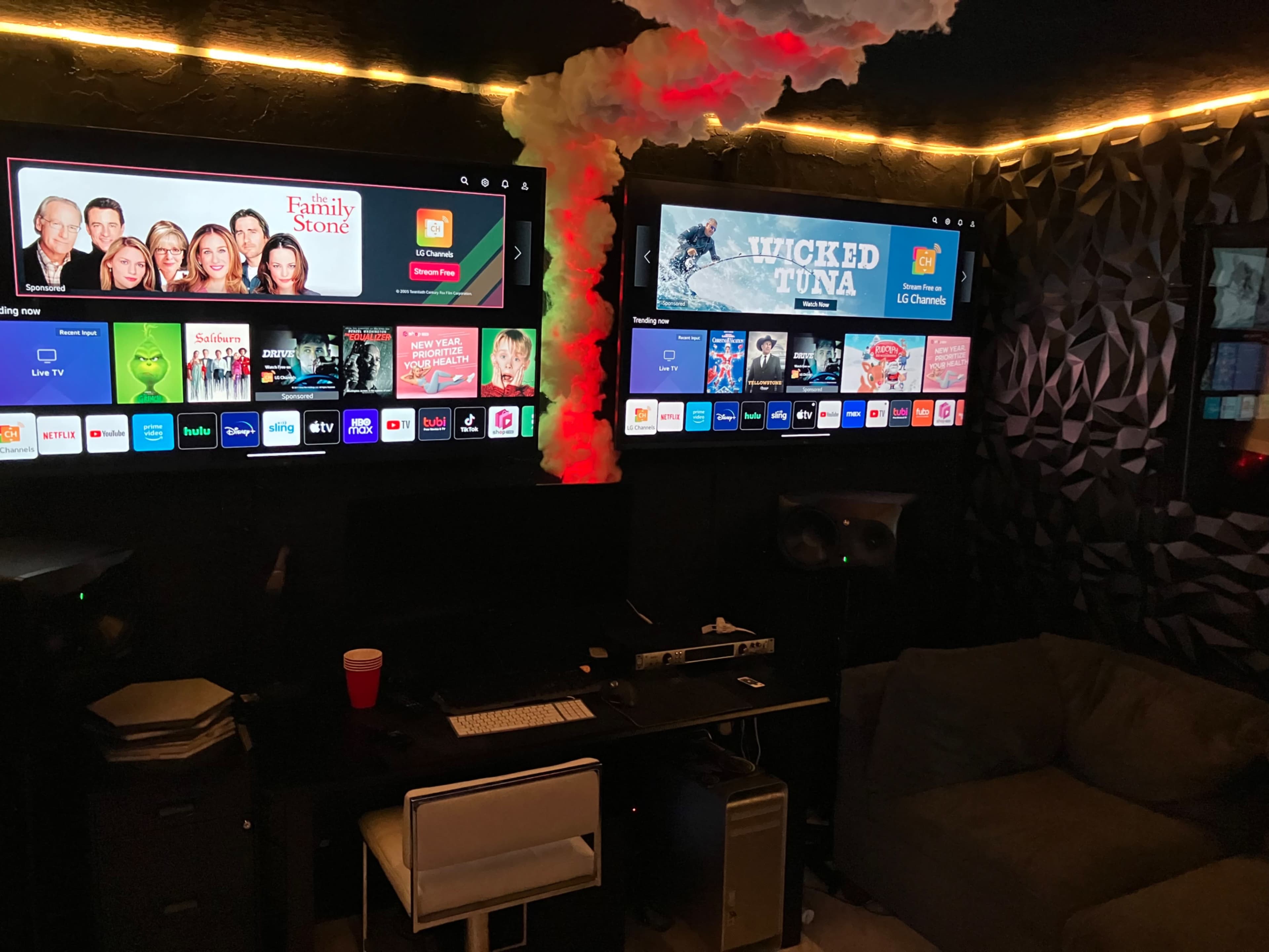 The image shows a dimly lit room with two large wall-mounted TVs displaying streaming content, a workspace with a desk and audio equipment, and a couch in the corner.