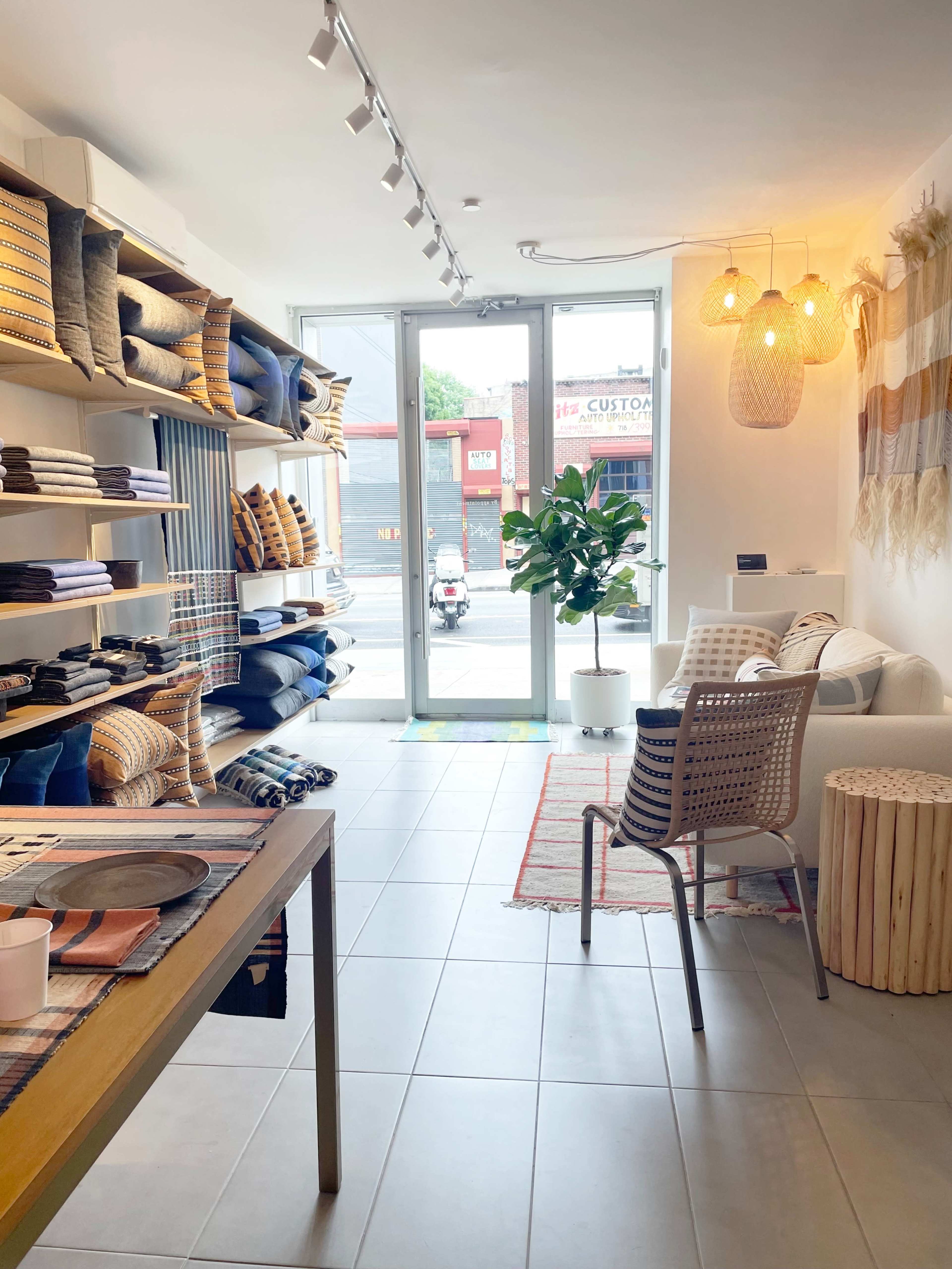 The image shows the interior of a modern home goods store with neatly organized shelves displaying textiles and a cozy seating area near large windows.