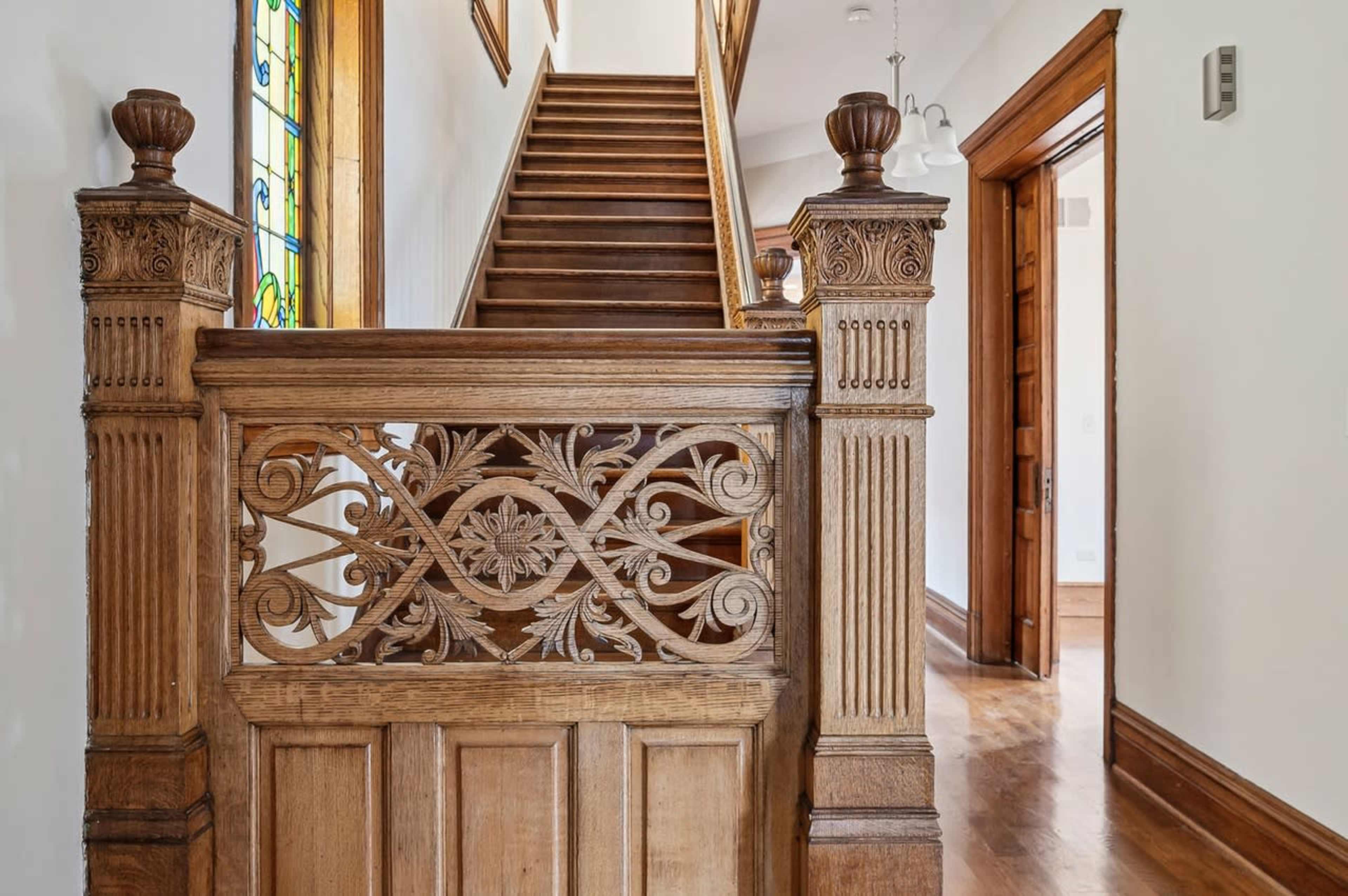 The image shows a wooden staircase with ornate balustrades leading to an upper floor, framed by a well-lit hallway.