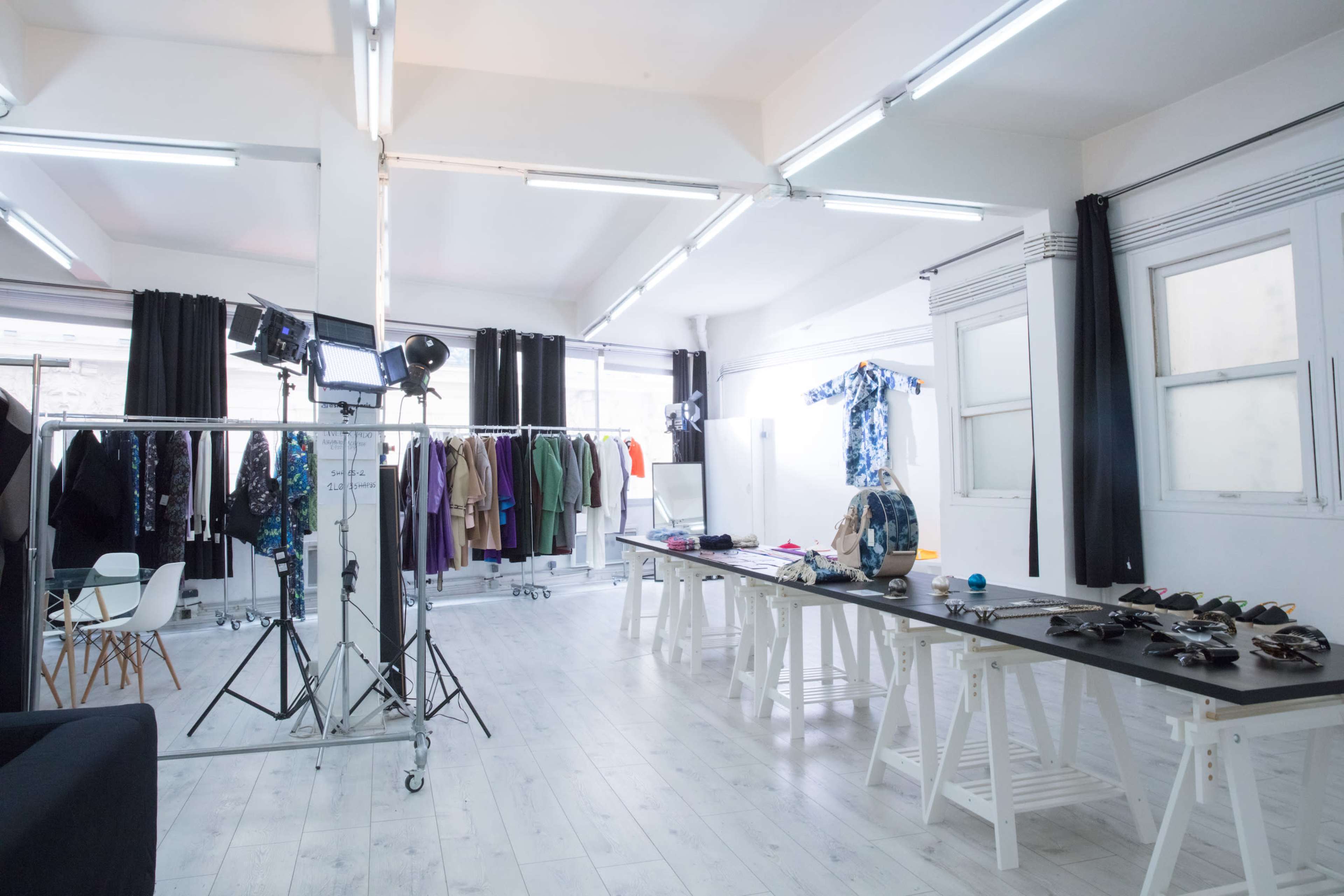 The image shows a well-lit fashion studio featuring racks of clothing, a table with various accessories, and a backdrop for photoshoots.