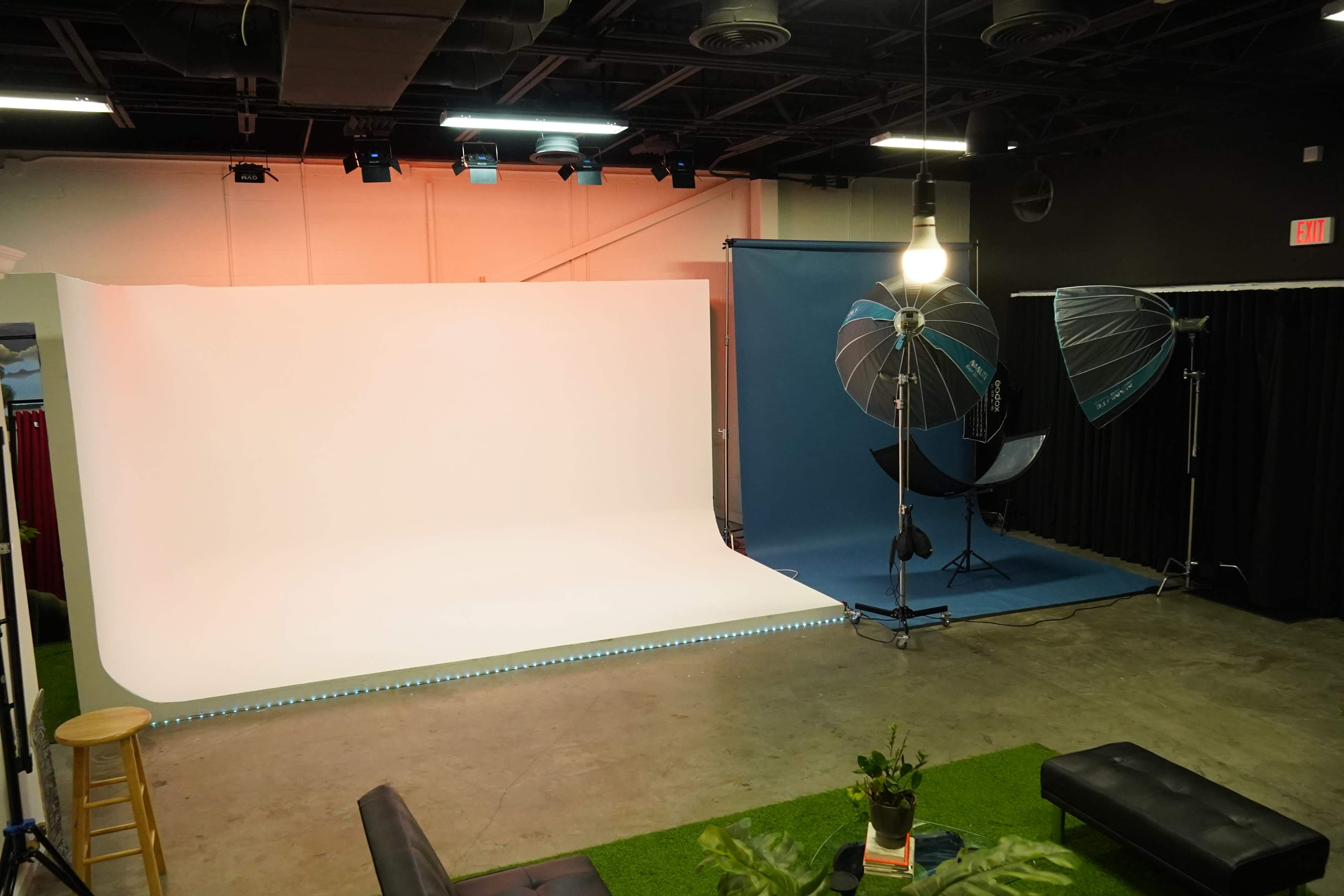 The image shows a photography studio with a white backdrop, a blue backdrop, studio lights, a stool, a black couch, and a small green area.
