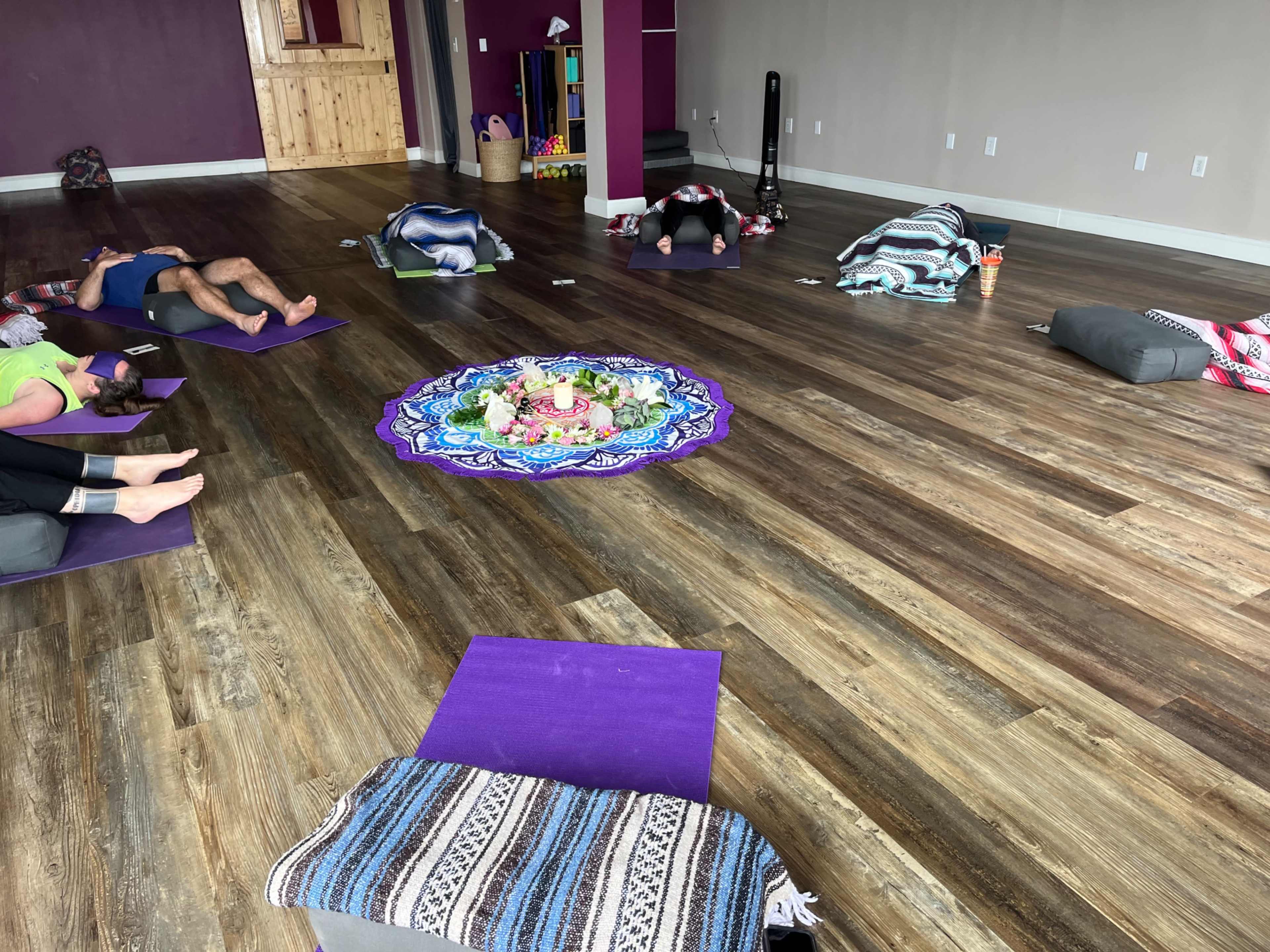 The image shows a yoga studio with several participants practicing on mats, surrounded by cushions and a colorful mandala in the center.