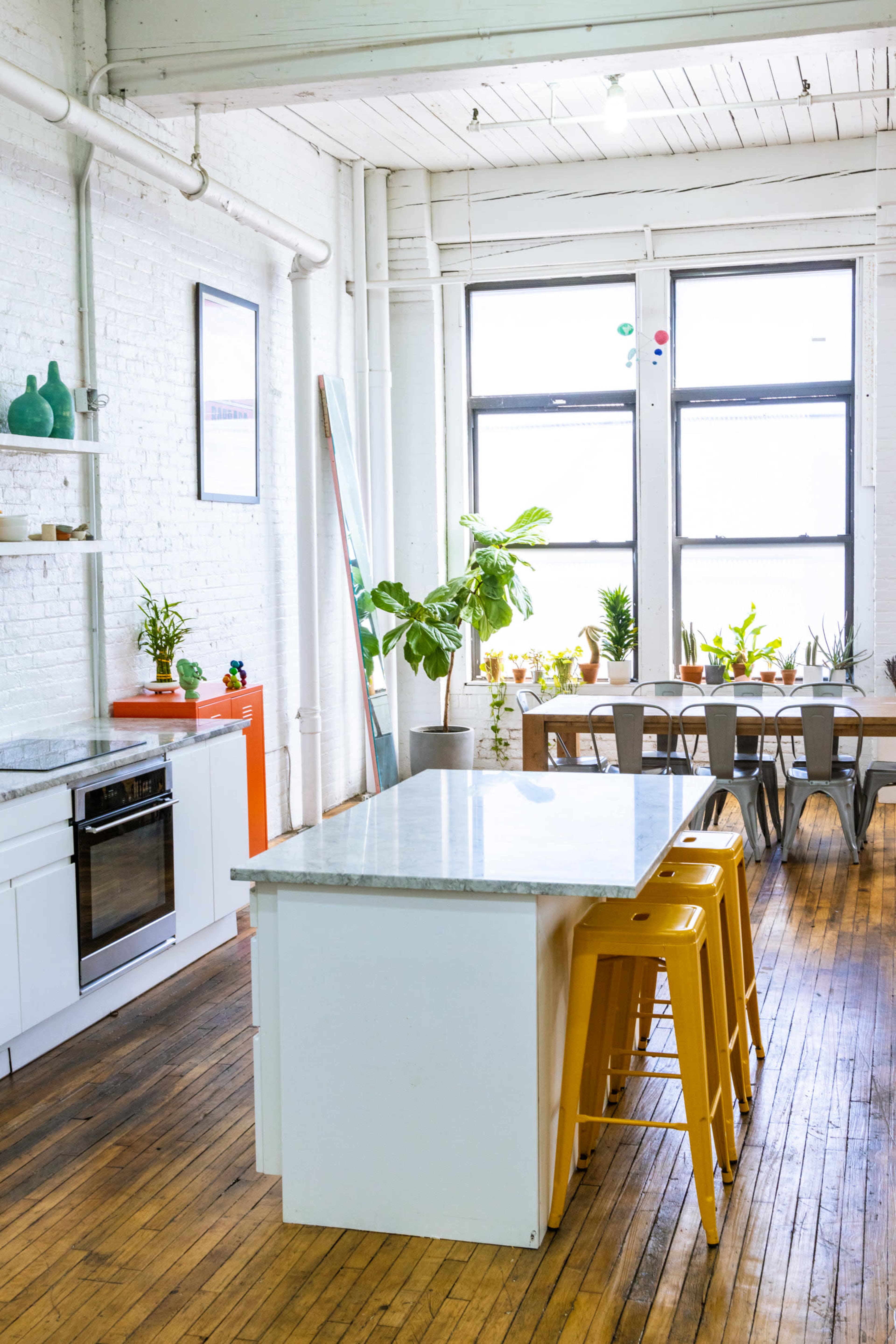 A spacious kitchen with a central island, yellow stools, and a dining area filled with plants and large windows.