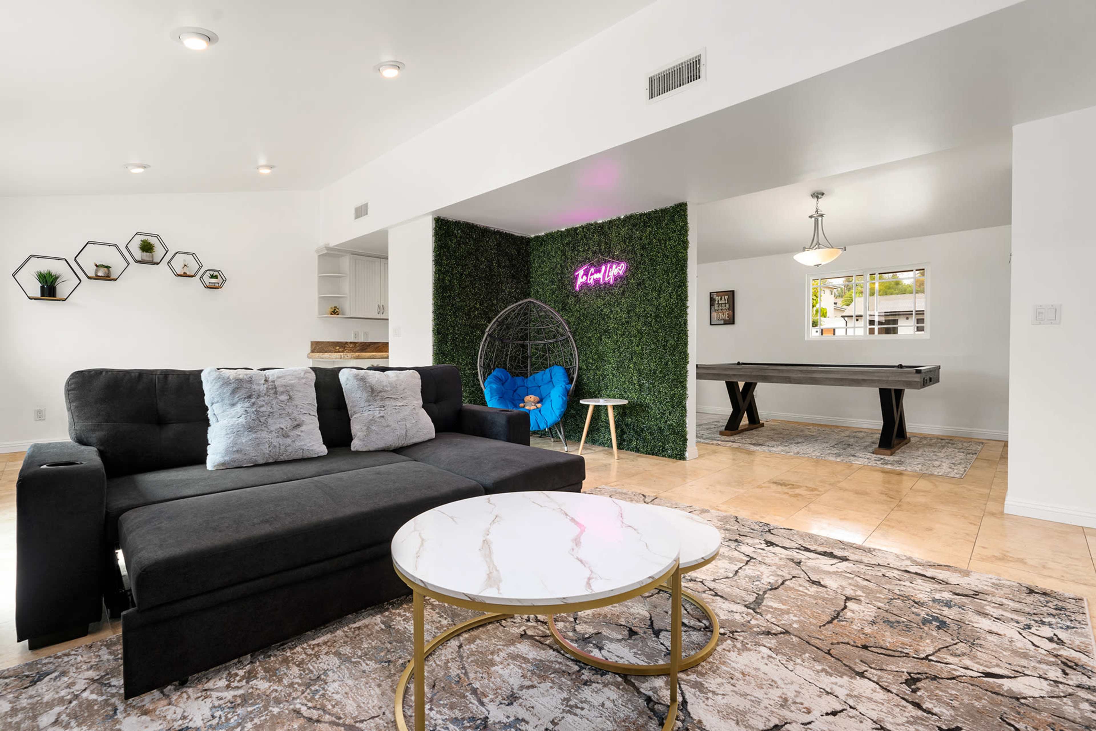 A modern living room with a black sofa, a circular coffee table, and greenery accents, leading to a game area with a pool table.