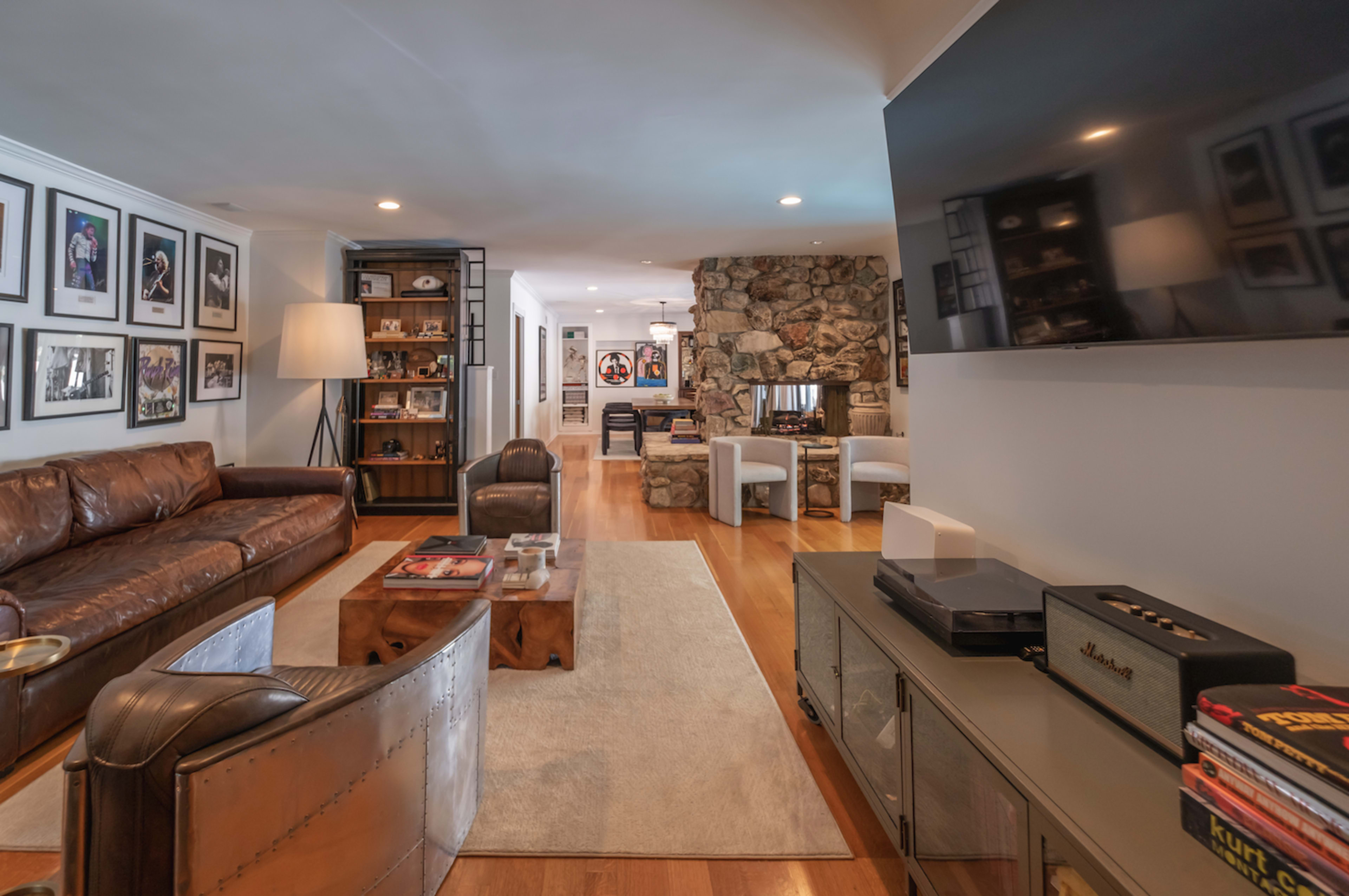 A living room featuring a brown leather sofa, a stone wall fireplace, and a flat-screen TV mounted on a white wall, with framed photos and bookshelves adding to the decor.