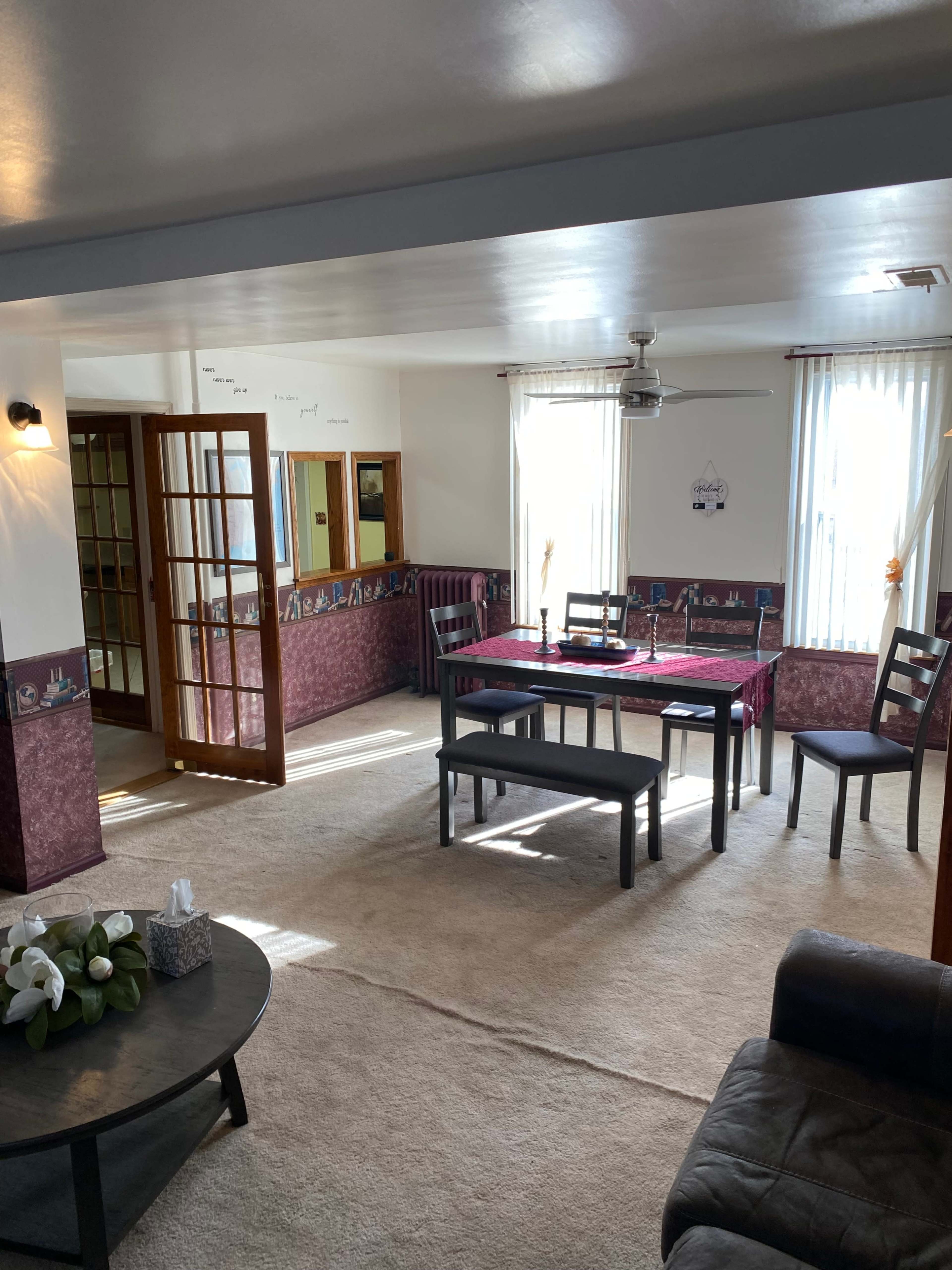 The image shows a living room with a carpeted floor, a table with chairs, a coffee table, and a set of double doors leading to another room.