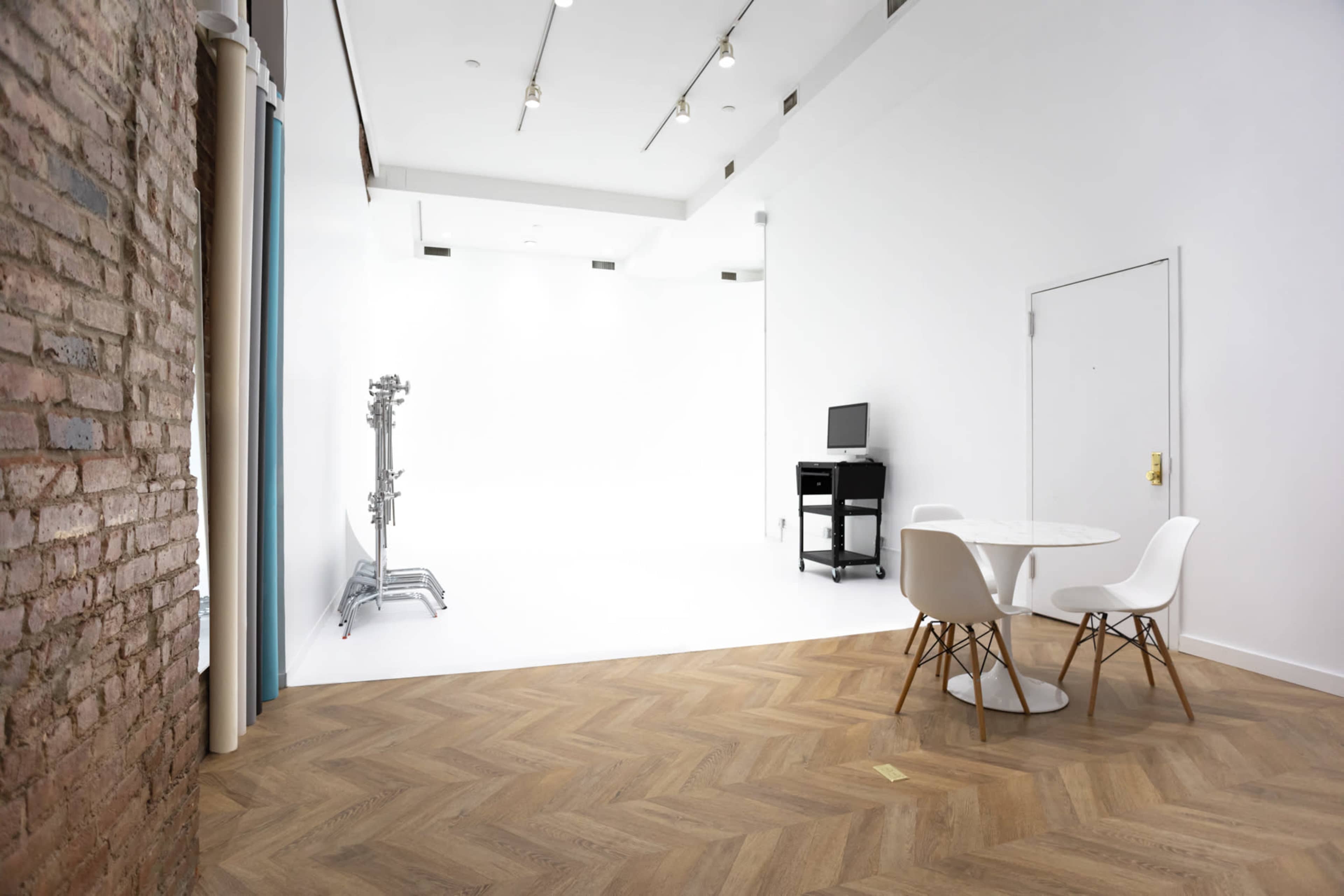 The image shows a spacious room with a wooden herringbone floor, a round white table and chairs, a computer station, and a backdrop stand against a white wall.