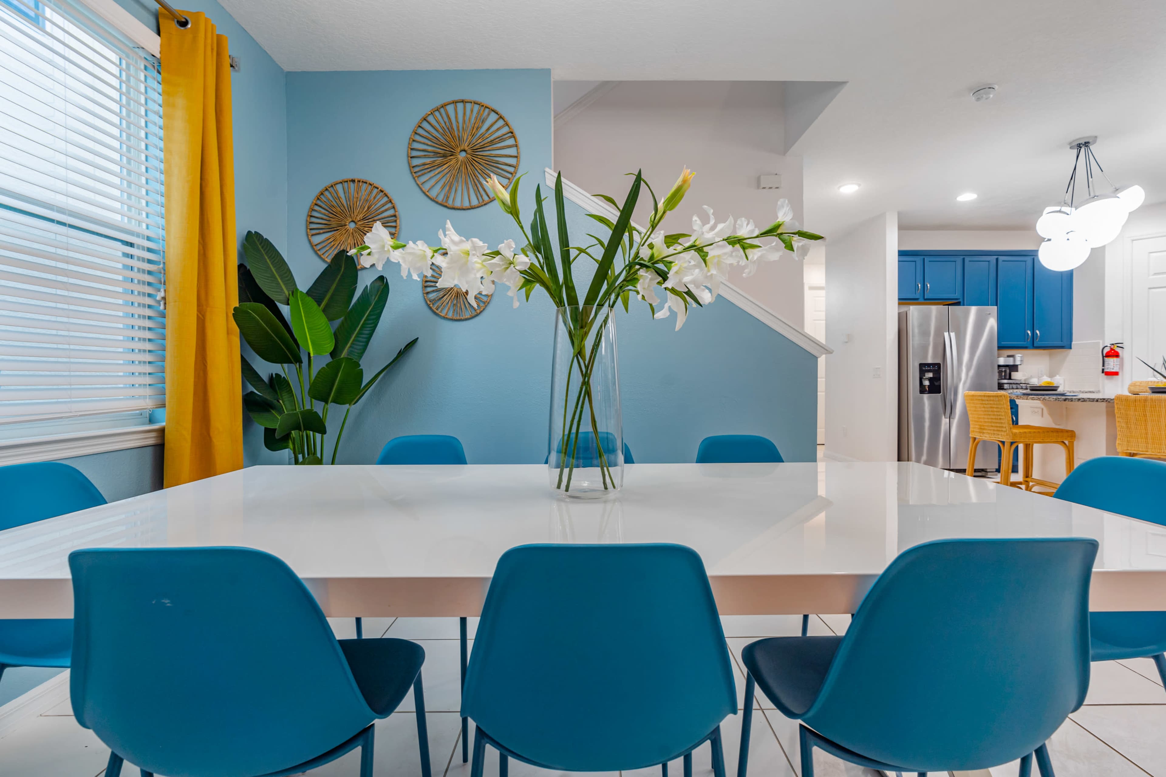 A modern dining area features a white table surrounded by blue chairs, with a vase of white flowers and a backdrop of blue walls and a yellow curtain.