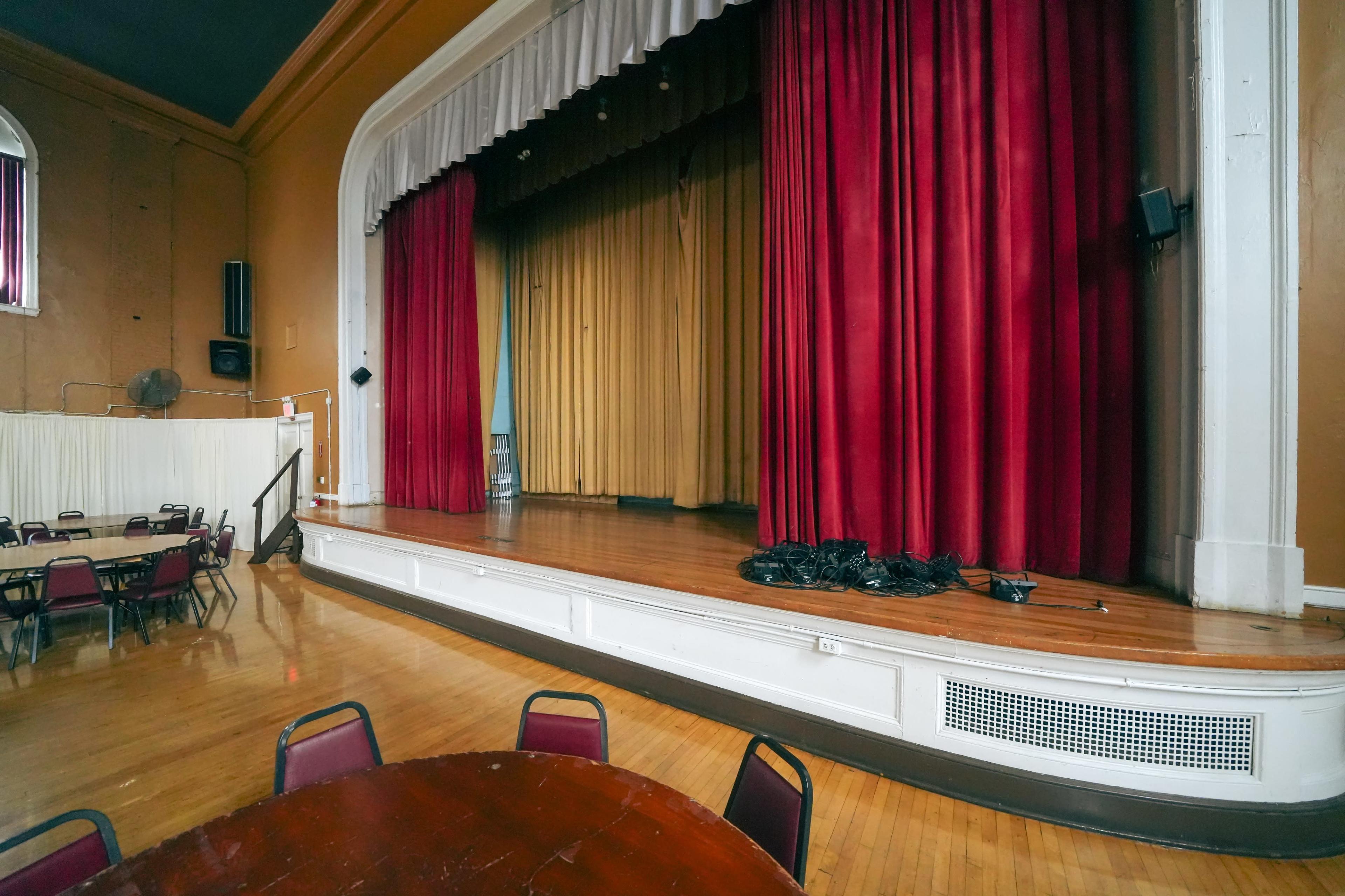 A stage in a theater with red and gold curtains, set against a wooden floor and a circular table with chairs in the foreground.